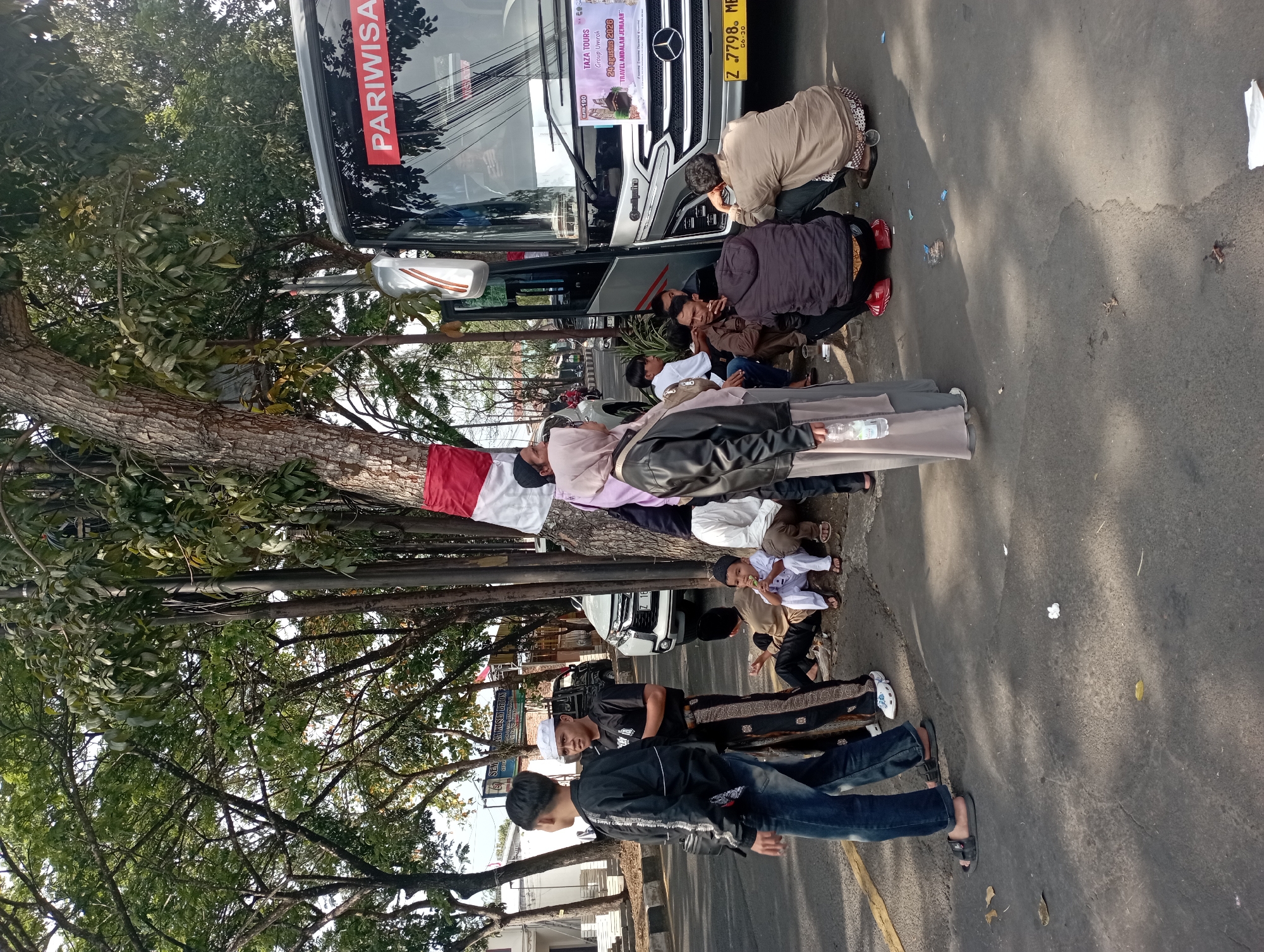 A group of people, some wearing Islamic attire like sarongs and peci hats, are gathered on a sidewalk next to a parked bus. The bus has "PARIWISATA" written on its side, indicating it is a tour bus. A banner on the bus windshield reads "TAZA TOURS GROUP UMROH 24 AGUSTUS 2023 TRAVEL AMBDALAN JEMAAH." Several individuals are seated on the ground, while others stand. The environment appears to be a street scene with trees, a road, and a building in the background. The license plate of the bus is visible and reads "Z 7798 MB". A red and white flag is tied to a tree. The location is identified as Tasikmalaya, Indonesia.