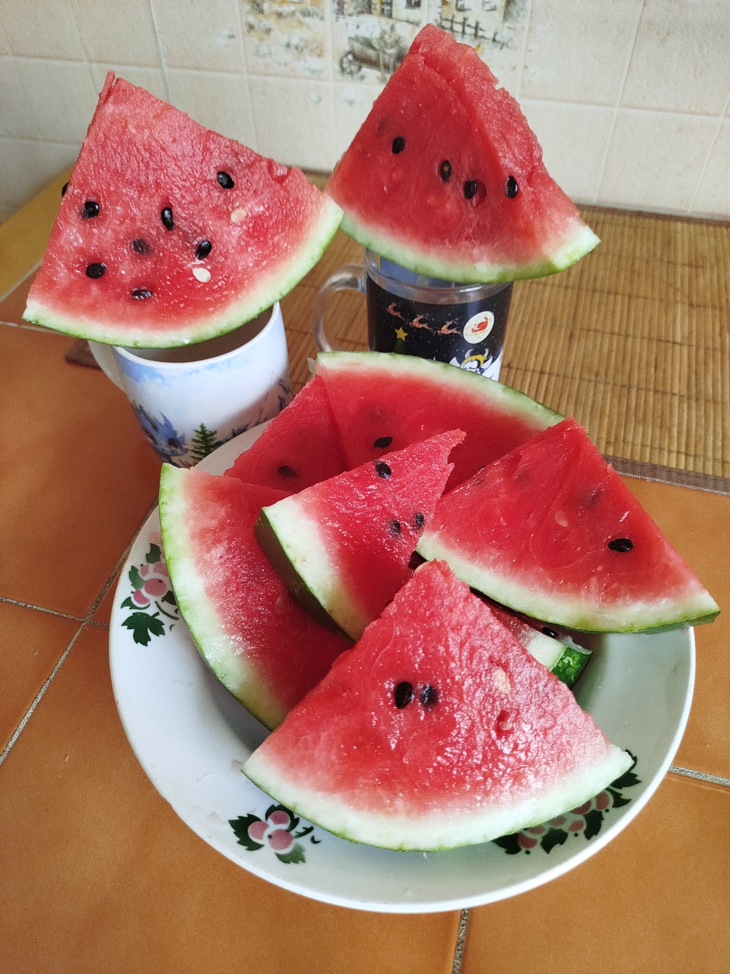 A plate of sliced watermelon sits on an orange tiled countertop. The plate is white with a floral pattern around the rim. There are several slices of watermelon on the plate, some stacked on top of each other. Two slices of watermelon are placed on top of mugs, one mug is white with blue and green imagery, the other is clear with black and white cartoon characters. The background consists of a tiled wall with a woven mat on the countertop.