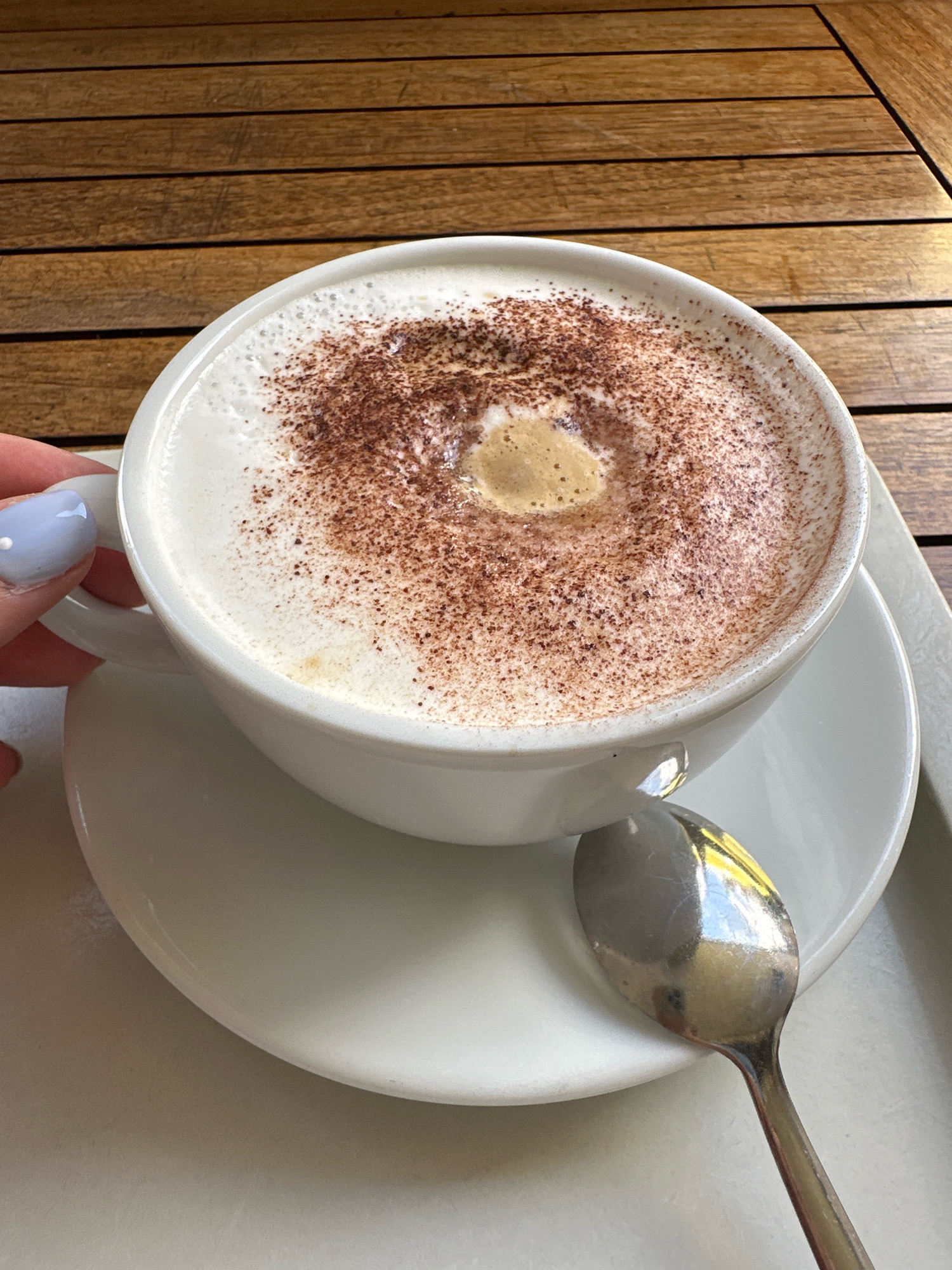 A white ceramic cup filled with cappuccino sits on a matching saucer. The coffee is topped with frothy milk dusted with cocoa powder, with a small, darker pool in the center. A silver spoon rests on the saucer to the right of the cup. A person's hand with light blue nail polish is visible holding the cup handle on the left side of the image. The setting appears to be outdoors, with a wooden slatted surface in the background, suggesting a cafe or restaurant patio. There is no specific information in the image to identify the city as Krefeld, Germany.
