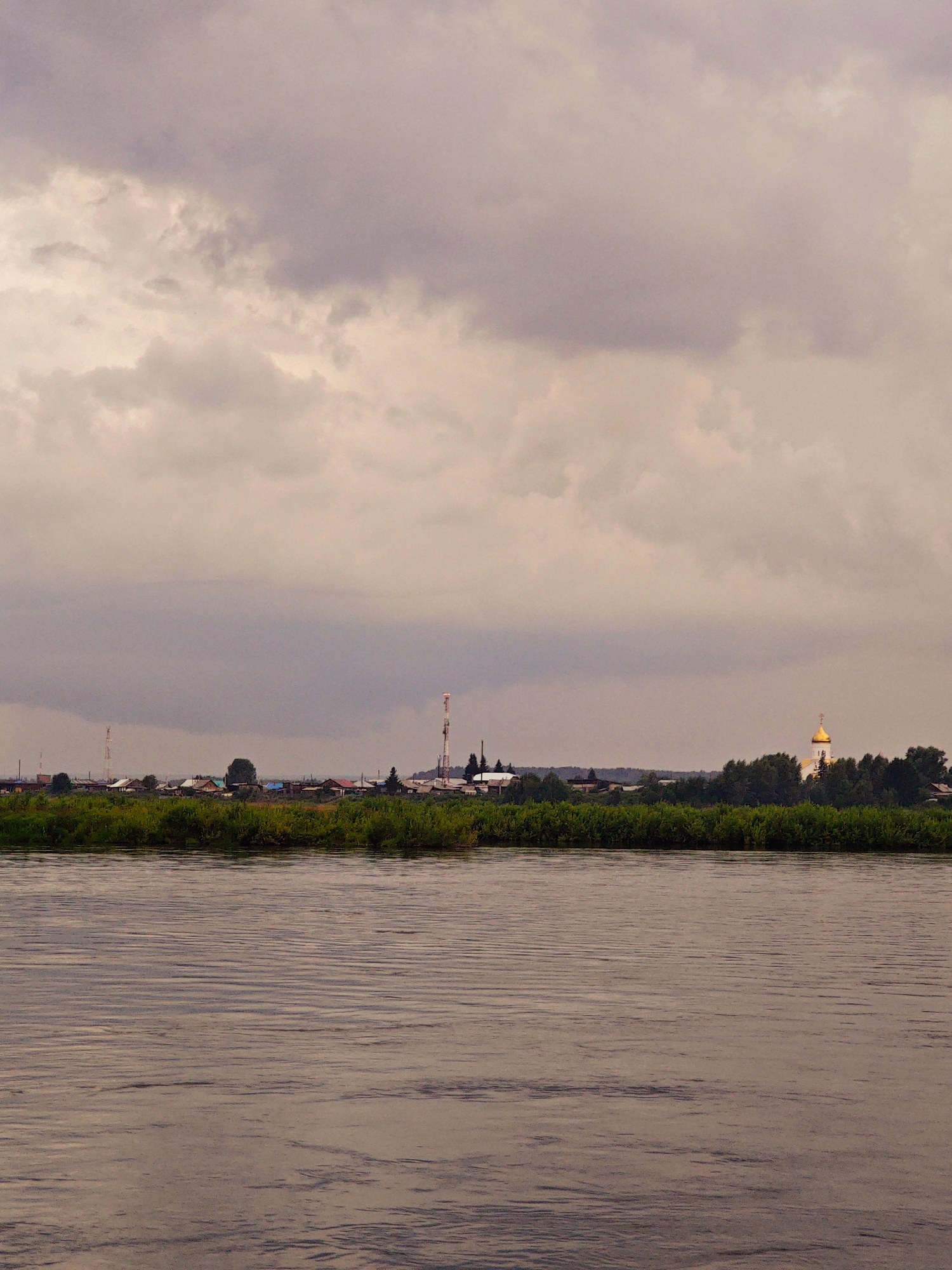 A wide, calm river fills the foreground of the image, its surface rippled by a gentle breeze. The water has a muted, brownish hue, reflecting the overcast sky. Beyond the river, a lush green embankment is covered in dense foliage. Nestled behind the vegetation is a small village. The buildings are a mix of houses with colorful roofs and some industrial structures, including two tall communication towers. To the right of the village, a golden-domed church stands out against the greenery, its spire catching the light. The sky above is filled with heavy, dark clouds, creating a somber, atmospheric mood. The overall scene suggests a quiet, rural setting, likely in the town of Kazachinskoe, Russia, given the architectural style of the church and the general landscape. No people are visible in the image.