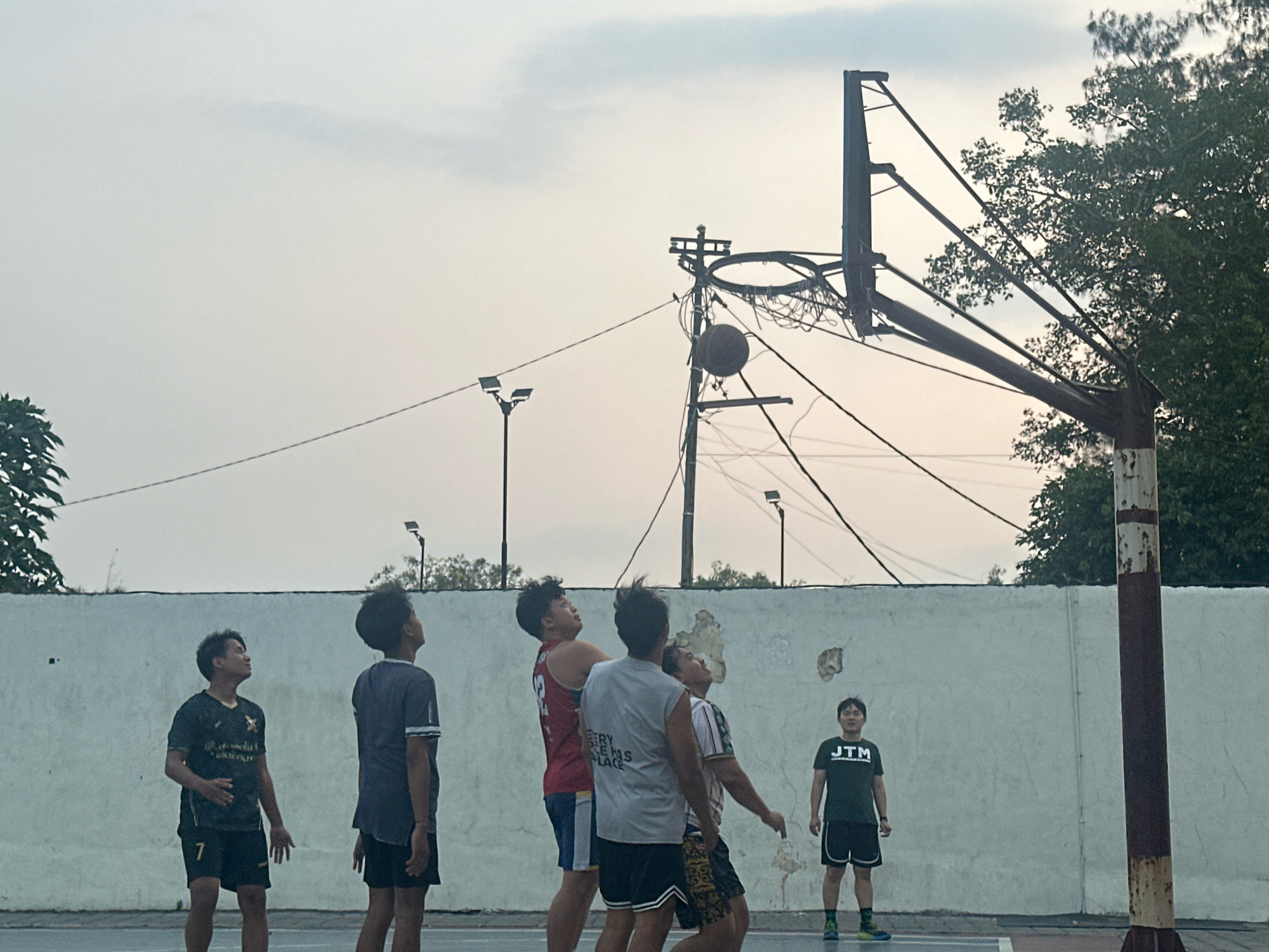 A group of six young men are playing basketball on an outdoor court. The court is enclosed by a white concrete wall. The sky is overcast and appears to be late afternoon or early evening. The basketball hoop is old and appears somewhat damaged, with wires hanging around it. The players are dressed in athletic wear and are in various stages of play, with some looking up at the ball. One player is positioned on the right side of the frame, watching the game. The image is taken from a low angle, looking up at the players and the hoop. The overall scene suggests a casual game of basketball being played in a local park or public recreational area. This image was taken in Mojokerto, Indonesia.