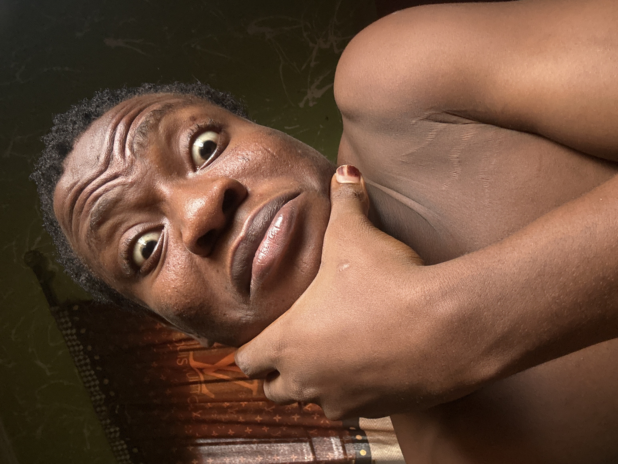 A person with dark skin and short, dark hair is positioned close to the camera. Their face is angled upwards, with wide eyes and a furrowed brow, giving the impression of surprise or concern. One hand, with a fingernail painted dark red, is placed against their cheek or jawline. The background includes a mottled wall in shades of green and brown. The setting appears to be indoors, near a window covered by a patterned curtain in brown and gold tones.