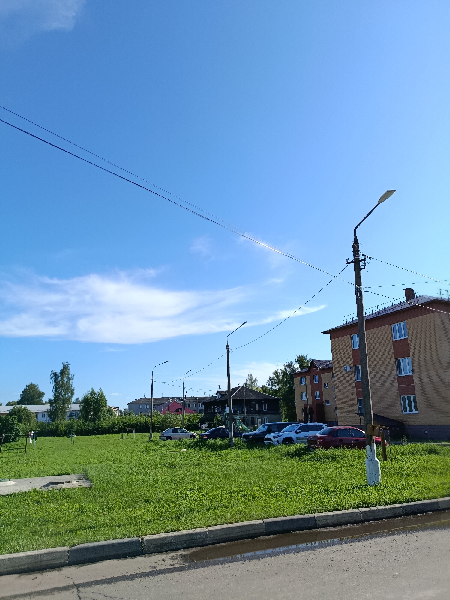 The image shows a street scene in the city of Vereya, Russia, on a clear day with a blue sky and scattered white clouds. In the foreground, there is a paved road with a concrete curb and a small puddle of water. Adjacent to the road is a grassy area with a few parked cars, including a red sedan and a white SUV. Beyond the grassy area, there are several buildings. To the right, a multi-story brick building with red-tiled roof is prominent, featuring multiple windows and a visible chimney. To the left of this building, other residential structures, some appearing to be older wooden houses, are visible amongst trees. A street light stands on a pole in the middle ground, and power lines are strung across the sky. The overall impression is of a residential area in a Russian town.