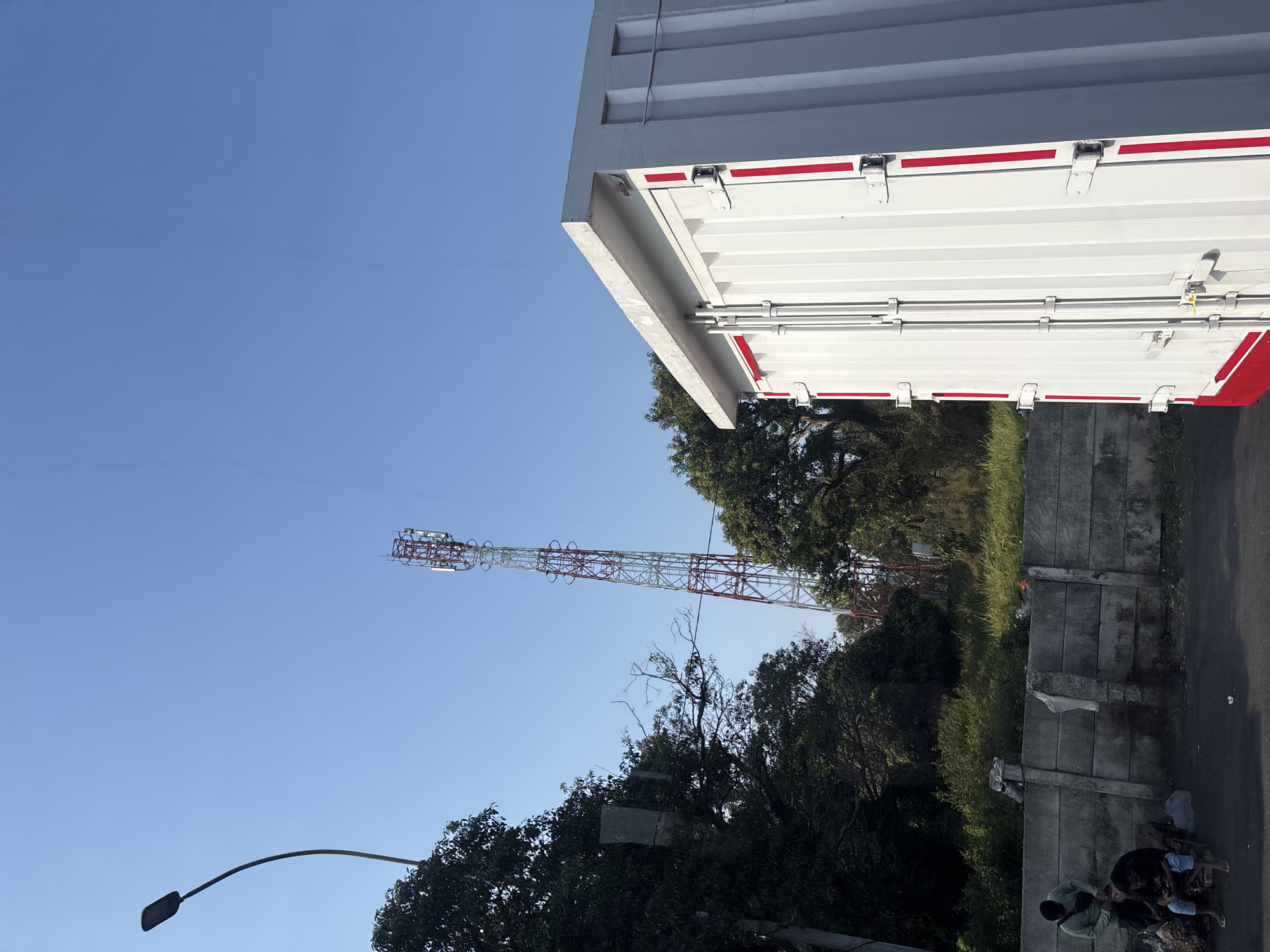 A cell tower stands tall against a clear blue sky in Purwakarta, Indonesia. In the foreground, a white shipping container with red accents is parked. To the left of the container, a concrete wall runs along the roadside. On the other side of the wall, two men are seated, with one appearing to be cutting the other's hair. Lush green trees and vegetation surround the area, with a streetlamp visible on the left side of the frame. The scene suggests an outdoor, possibly semi-urban environment.