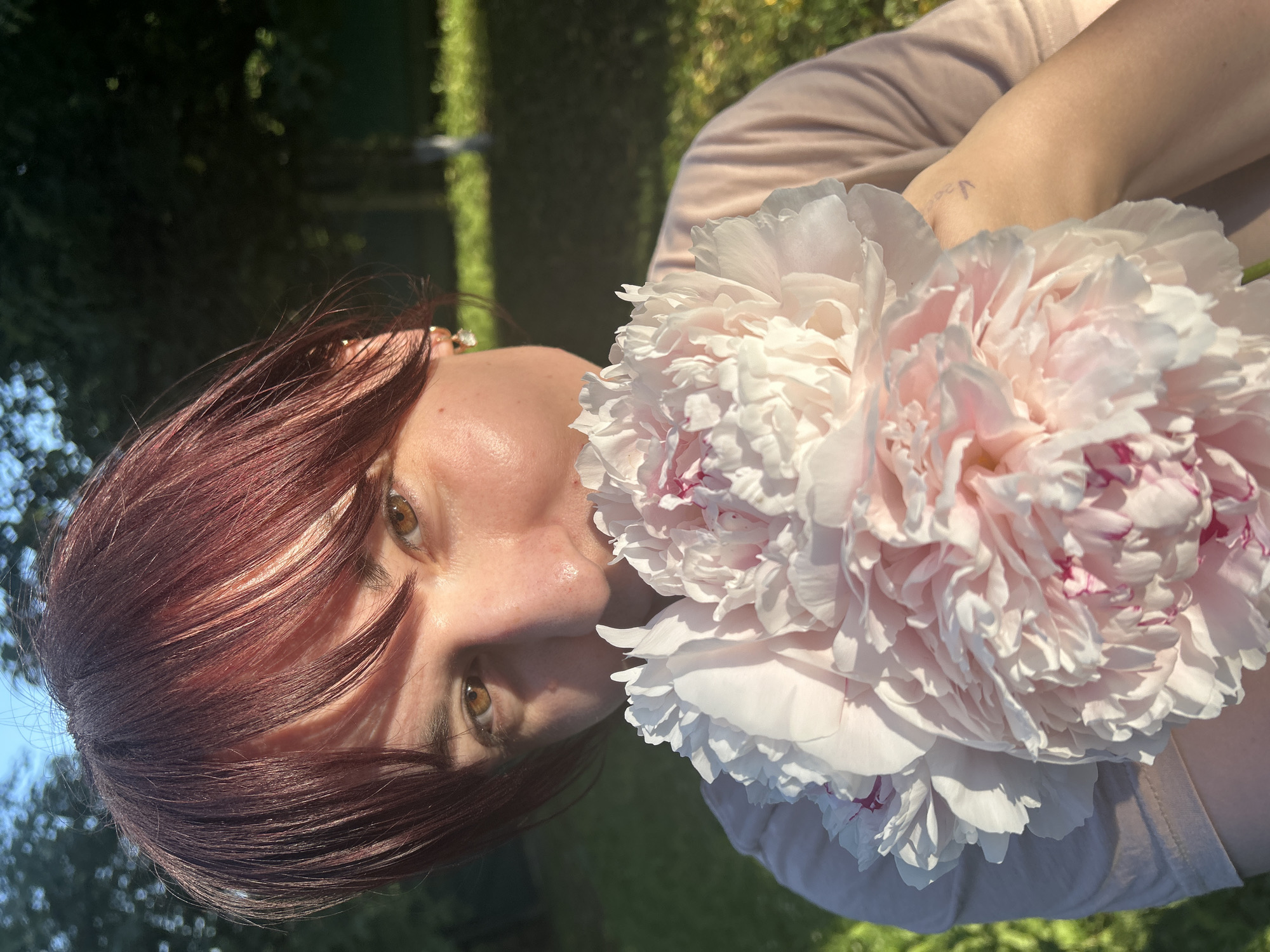 A close-up, vertically oriented medium shot captures a person with auburn hair holding a large, pale pink peony flower in what appears to be an outdoor setting in Foky, Russia. The person's face is partially obscured by their hair and the flower, but their eyes are visible, looking towards the camera with a soft, gentle gaze. The flower is the dominant object in the frame, its delicate petals a mix of white and soft pink, with hints of darker pink in the center.
The background is out of focus, showing hints of green foliage and what might be a garden or park setting. The lighting suggests it is daytime, with sunlight casting shadows and highlighting the textures of the flower and the person's skin. The overall mood is serene and peaceful, evoked by the softness of the flower, the gentle expression, and the natural light. There is no visible text in the image.