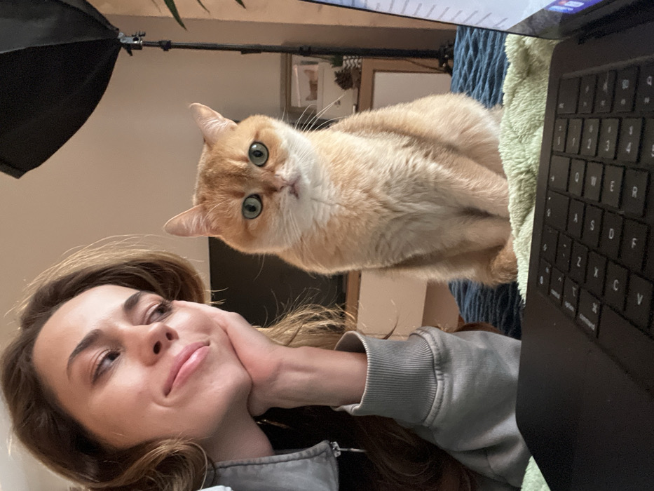 The scene is an indoor setting in Bar, Montenegro, featuring a female and a cat. A fair-skinned female, with light brown hair and wearing a grey hooded garment, is positioned on the left, resting her chin on her right hand while looking downwards and left with a subtle smile. Adjacent to her, a short-haired orange and white cat with green eyes sits erect, directly facing the camera. Both subjects are situated on a light green textured blanket, which overlays a darker teal-colored blanket. A black laptop's keyboard and a partial view of its illuminated screen are present in the foreground on the right. The blurred background reveals a black studio light on a stand to the right of the cat, a wall-mounted television over a white cabinet, and a light-toned shelf containing items such as pinecones and framed objects. A green-leaved plant is partially visible on the extreme right.