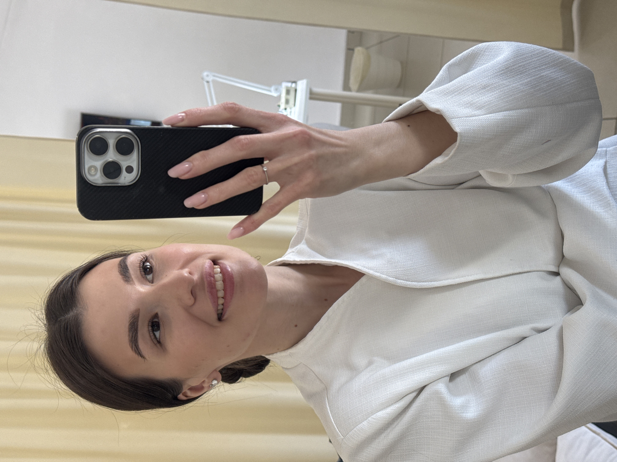 A woman is taking a selfie with a black smartphone, presumably an iPhone based on the three camera lenses visible on the back. She is wearing a white garment with puffed sleeves. A silver ring adorns one of her fingers. The background includes a white wall, a curtain, and an adjustable lamp. Given the prompt, this scene may be located in Chaikovsky, Russia, but the image itself offers no definitive confirmation of this location.