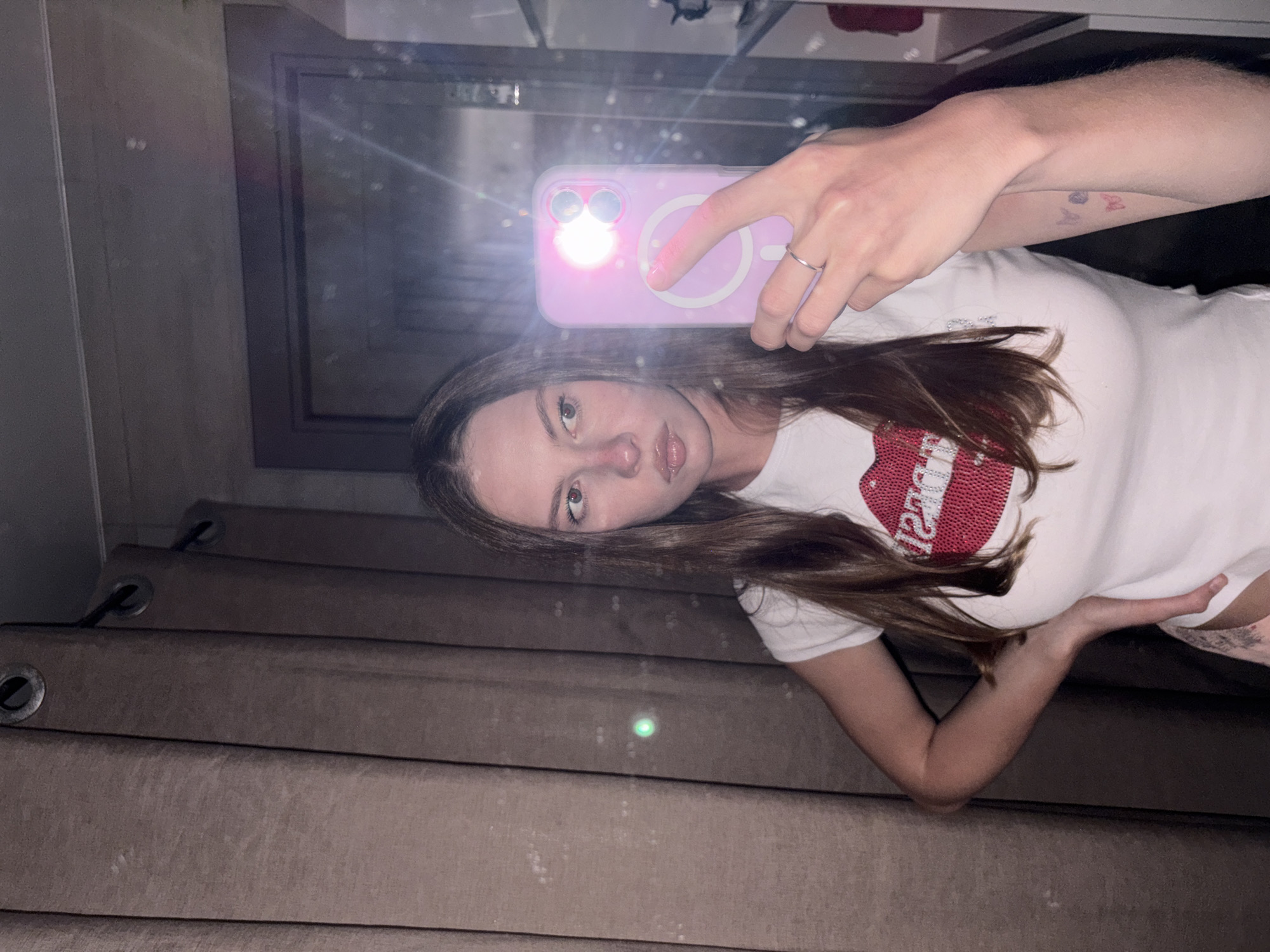This image depicts a young, light-skinned female taking a mirror selfie indoors in the city of Perm, Russia. She has long, straight brown hair and is looking to her right with a neutral expression, her lips slightly parted. She wears a white, short-sleeved t-shirt featuring a large, red, glittery lip design on the chest with the mirrored word "DESIRE" visible. A light-colored patterned garment is partially visible below her t-shirt on her left side. She holds a light purple iPhone, equipped with a circular white MagSafe ring, in her right hand, with a silver ring on her right ring finger. The phone's flash is active, producing a bright glow and lens flares across the mirror's surface. Her left hand rests on her hip, revealing small, multi-colored butterfly tattoos on her left forearm. The background, reflected in the mirror, shows dark brown curtains with metal grommets on the left, displaying a faint green laser dot and several small dust particles or smudges on the mirror itself. The right side of the background reveals dark paneling or a wall, with shelves or a counter further right, containing indistinct objects.