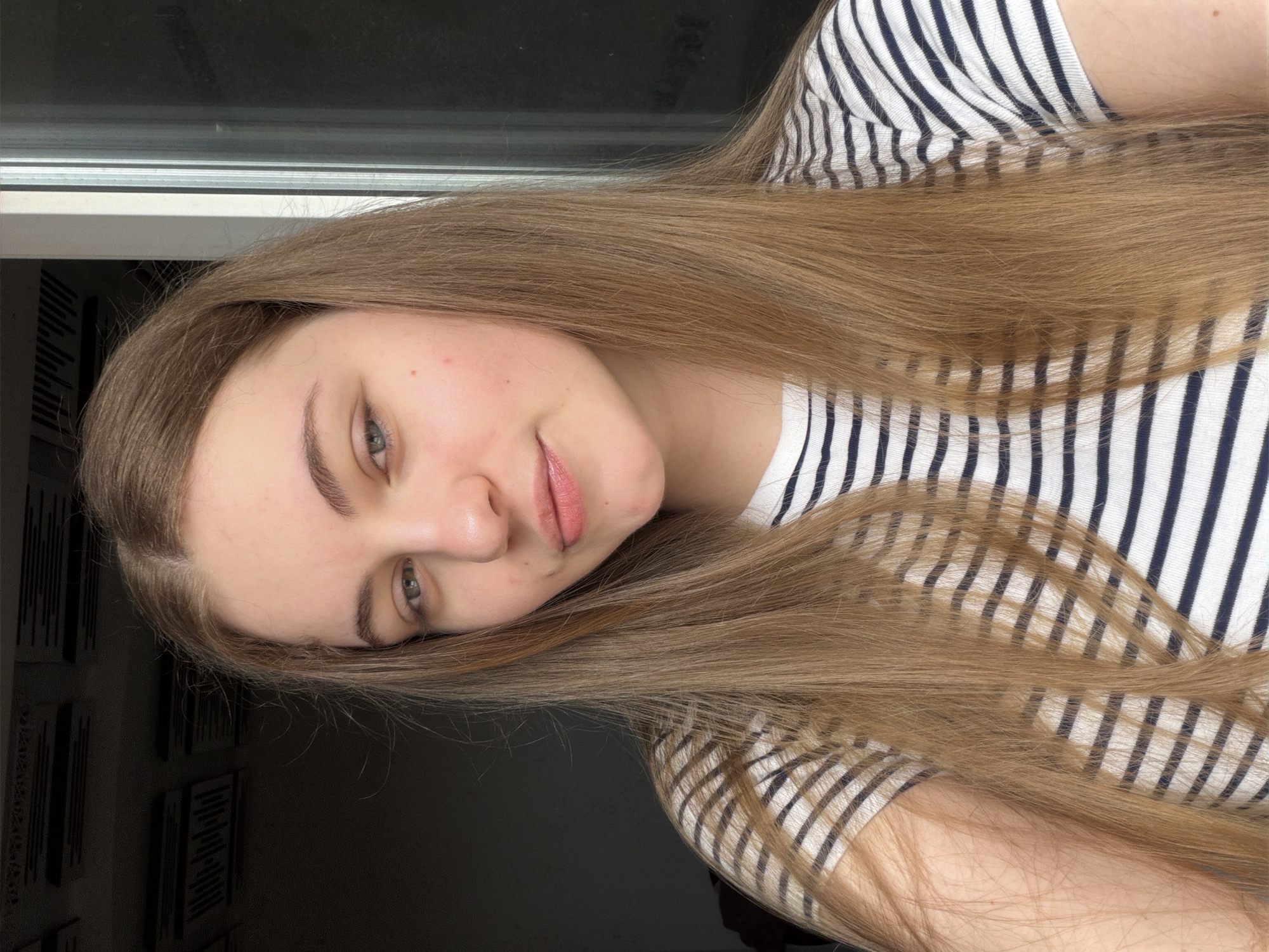 A young woman with long, straight, light brown hair and light blue eyes is shown in a close-up, waist-up shot. She is wearing a striped t-shirt with horizontal navy and white stripes. Her skin is fair with some light freckling. She is looking directly at the camera with a neutral expression. The background consists of a dark wall with some horizontal elements, possibly shelves or acoustic panels, on the left, and a window with vertical blinds on the right. The lighting suggests it is daytime, possibly indoors near a window. The image appears to be a selfie taken in an indoor environment, likely a residential setting in Dnipro, Ukraine, given the context.