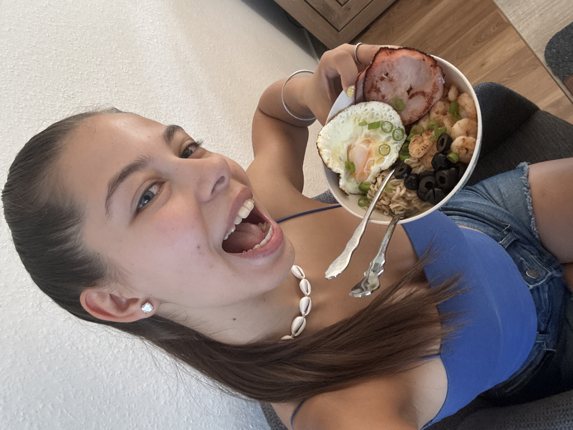 A young woman with dark brown hair pulled back in a ponytail, wearing a blue tank top and denim shorts, holds a bowl of food. The bowl contains noodles, a fried egg, shrimp, black olives, a slice of ham, and chopped green onions. She is looking at the camera with her mouth open, appearing excited to eat. She is wearing a seashell necklace and a silver bracelet on her right wrist, and a ring on her left ring finger. The background is a plain white wall. The image does not provide any information about the city of Bruchköbel, Germany.
