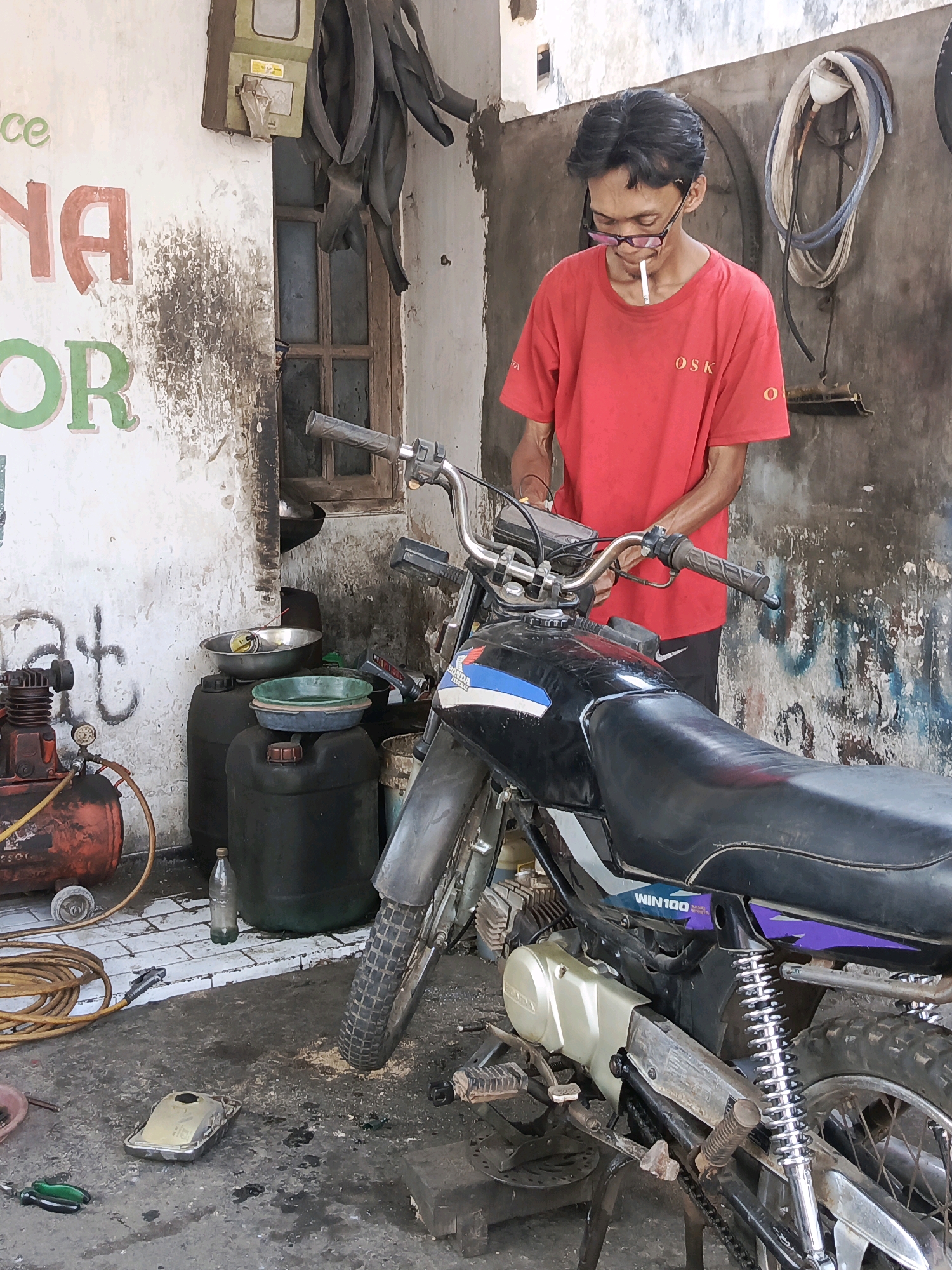 In Semanding, Indonesia, a man with black hair and glasses is standing next to a black motorcycle in what appears to be a motorcycle repair shop. He is wearing a red t-shirt and black pants, and a cigarette is lit in his mouth. The motorcycle is black with blue and white decals. In the background, there is a concrete wall with some graffiti, a window, and various tools and supplies related to motorcycle repair, including an air compressor, large plastic barrels, and hoses. The floor is concrete and appears to be dirty. The overall environment suggests a functional, working space for vehicle maintenance.