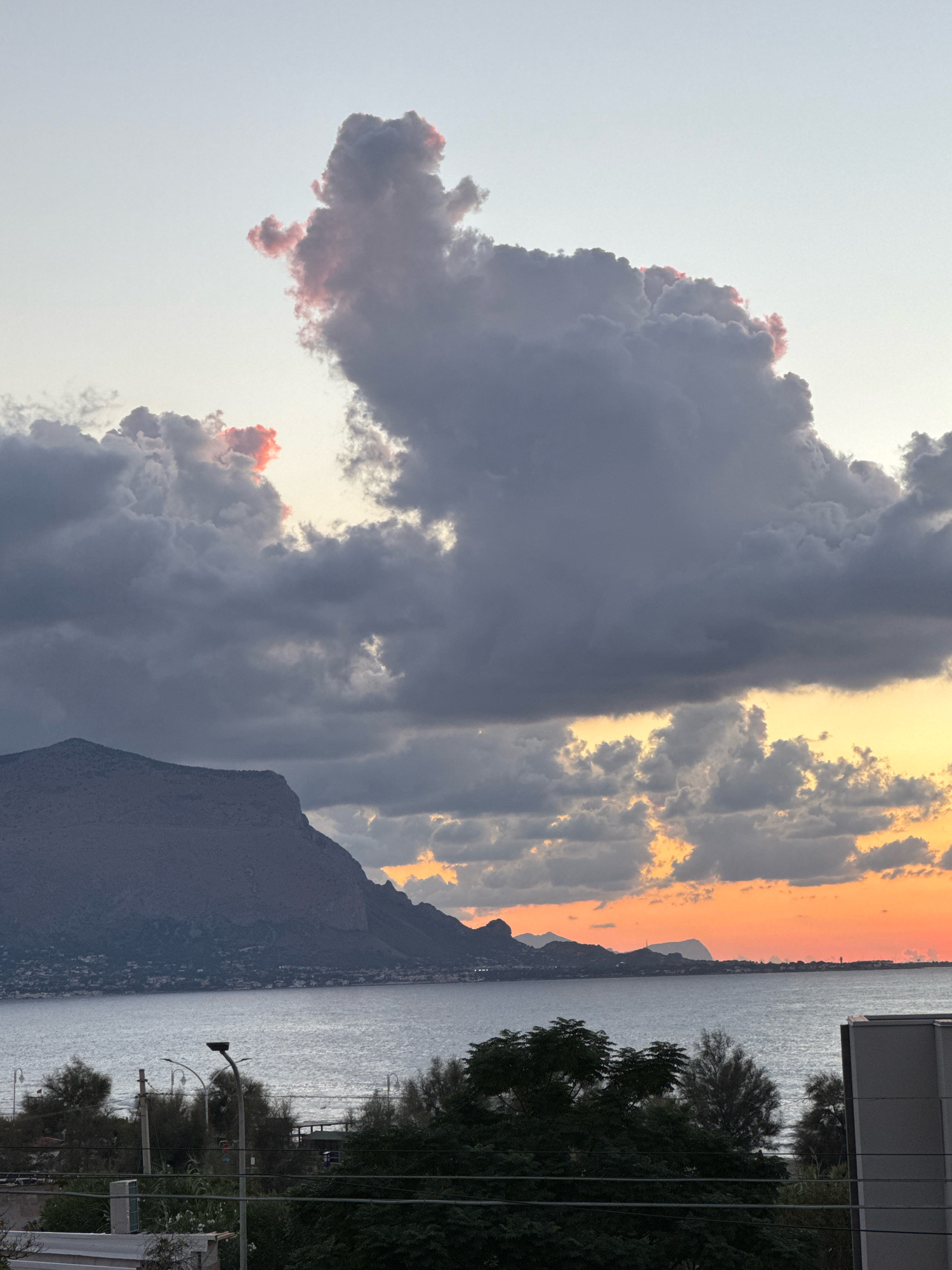 A landscape scene depicts the coast of Capaci, Italy, at sunset. A large mountain range dominates the left side of the frame, with a body of water in the foreground. Dark, ominous clouds fill the sky, with hints of pink and orange light breaking through on the right side, indicative of the setting sun. The water is calm, reflecting the twilight sky. In the foreground, trees and vegetation are visible, along with utility poles and streetlights, suggesting an urban or semi-urban setting. The overall atmosphere is dramatic and serene, with the natural beauty of the landscape juxtaposed with man-made elements.