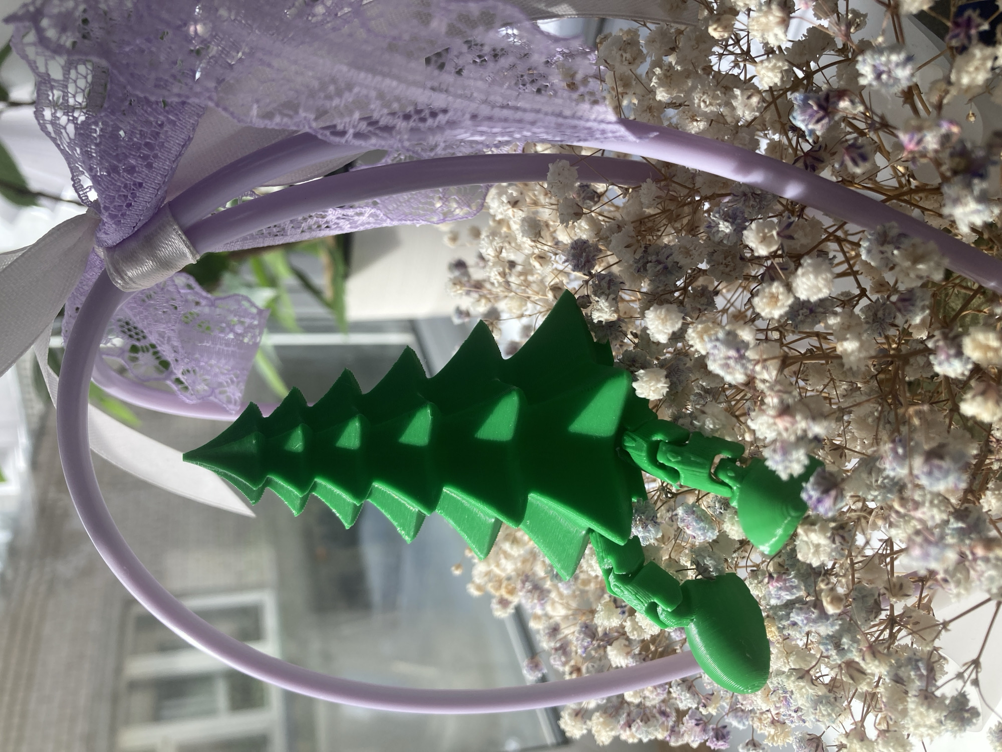 The image presents a still life composition featuring a green, articulated 3D-printed Christmas tree figure, a light purple hoop, and dried flowers, set on a reflective surface. The central object is a bright green, segmented Christmas tree-shaped toy, displaying distinct 3D print lines. It has movable joints, with two bulbous, limb-like appendages extending to the right, suggesting a playful design.
A delicate light purple lace fabric, secured by a thin white ribbon, is draped in the upper left of the frame. This lace partially covers a smooth, light purple hoop that curves across the lower and middle sections of the image.
The background is dominated by a dense cluster of dried baby's breath flowers (Gypsophila), predominantly white with subtle faded purple and blue tones. These flowers provide a soft, textural contrast to the geometric lines of the artificial tree and hoop.
All items are placed on a highly reflective surface, likely glass or polished material, which creates clear mirrored images of the objects, particularly the purple hoop. The scene is illuminated by bright, natural daylight, indicating an indoor setting during daytime. Blurred reflections in the background hint at windows or other interior structures. No specific activity is taking place, and no text is visible. This scene is located in Chaikovsky, Russia.
