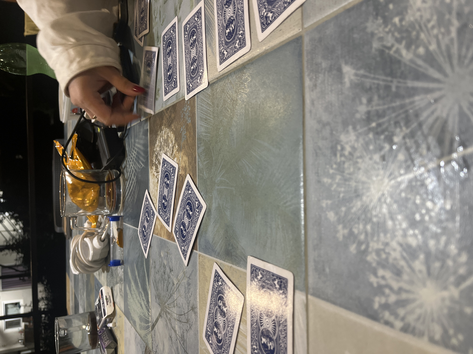 In an indoor setting, likely a home, a close-up shot focuses on a table covered with a patterned surface. Several playing cards are spread out on the table, some face down and others face up. A person's hand, with red nail polish, is reaching towards one of the cards. To the left of the hand, a glass mug filled with a golden liquid and a yellow-bagged snack are visible. Further to the left, another glass with a dark liquid and a pack of cigarettes are present. A white coiled object, possibly a charger, and a small blue and clear object, perhaps a timer, are also on the table. In the background, a blurred view of a building with windows and trees is discernible. The lighting suggests it is nighttime.