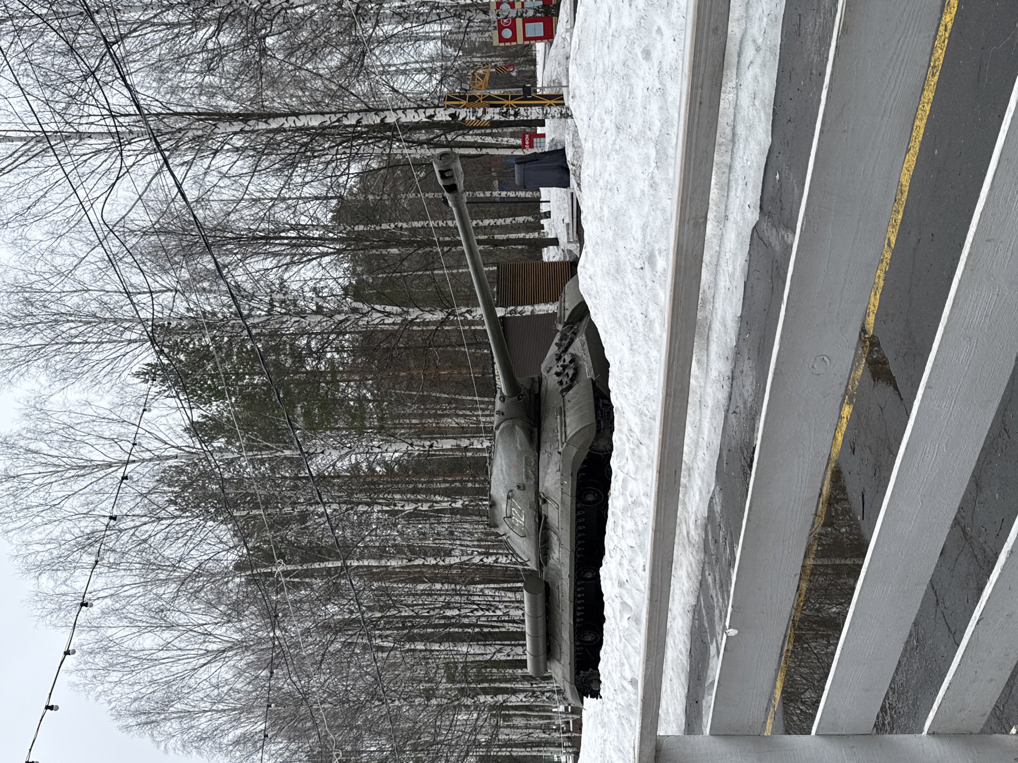 An outdoor, daytime scene shows a Soviet/Russian T-54 or T-55 tank in Izhevsk, Russia. The tank is painted in a dark green/grey color. It is positioned in a winter landscape with snow on the ground. The tank is facing left in the image. Behind the tank, there are leafless birch trees. In the upper background, there are structures, possibly a playground. The foreground features a series of white, horizontal wooden planks with gray or yellow lines visible between them, possibly part of a viewing platform or structure. The sky is overcast.