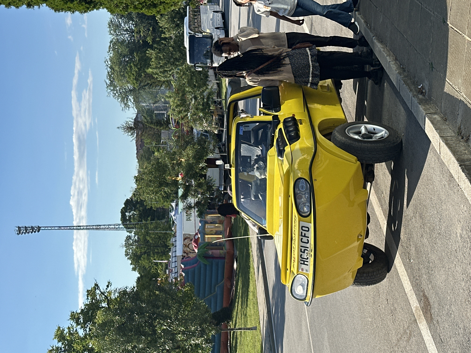 A bright yellow amphibious vehicle, a "boat car" according to its license plate which reads "HC51 CFO UK", is parked at the curb on a paved road. The vehicle has a distinctive front end with round headlights and a broad bumper. Several people are gathered near the vehicle, some looking at it. In the background, an inflatable bouncy castle is set up on a grassy area, with tents and a tall light tower also visible. The scene appears to be a public outdoor event or fair, possibly in a park or open space within the city of Gorna Oryahovitsa, Bulgaria, as indicated by the overall atmosphere and the presence of event infrastructure. The sky is clear blue with a few white clouds, and sunlight illuminates the scene.