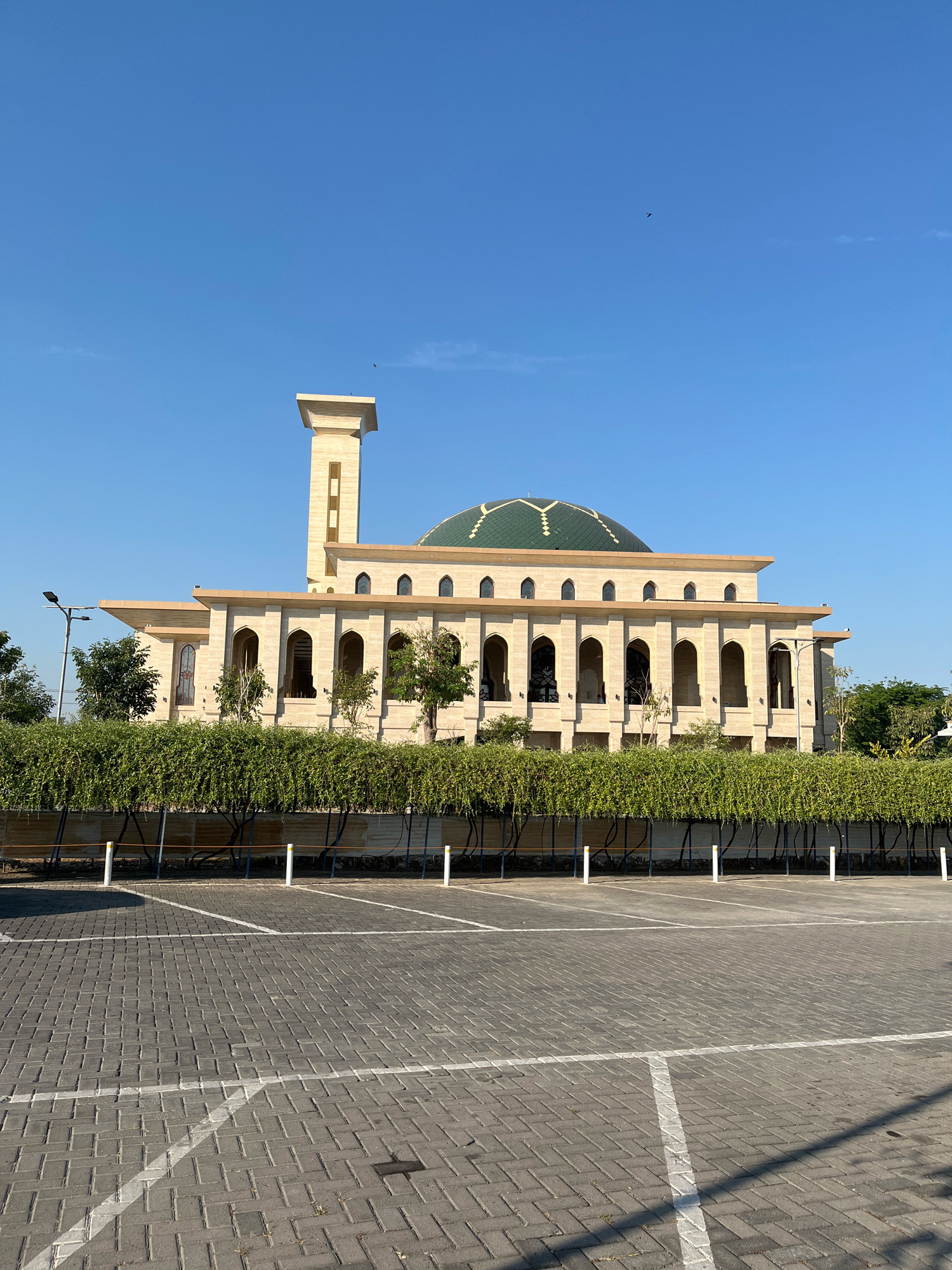 The image shows a large, light-colored building with a green dome and a tall minaret, likely a mosque, set against a clear blue sky. The building is ornate, featuring multiple arched windows and decorative elements. In front of the building is a neatly trimmed hedge, behind which is a paved parking lot with white parking lines. The ground of the parking lot is made of interlocking gray bricks. A street lamp is visible to the left of the building. The scene appears to be in a well-maintained outdoor public space. There are no people or animals visible in the image. The city of Dapur, Indonesia is not depicted in this image.
