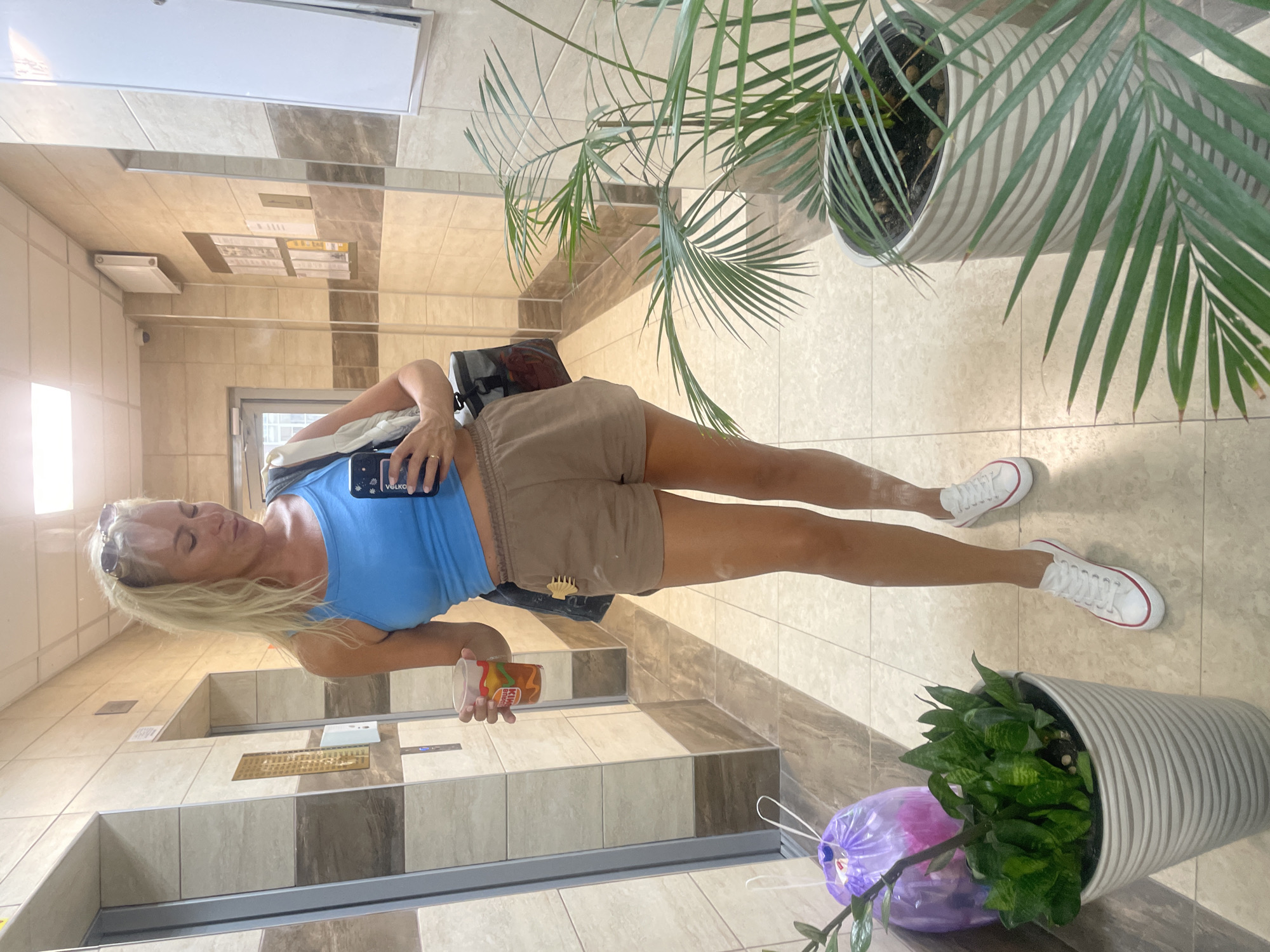 A woman with blonde hair pulled back in a ponytail, wearing sunglasses pushed up on her head, is taking a mirror selfie in a tiled hallway with elevators. She is wearing a blue tank top, brown athletic shorts, and white low-top sneakers with red accents. In her left hand, she holds a clear plastic cup with a beverage and ice. A dark gray shoulder bag is slung over her right shoulder. To her left, near an elevator door, is a large gray planter with a snake plant and a purple plastic bag. To her right, a large white pot contains a palm plant. The environment suggests a residential building in Novorossiysk, Russia, based on the tiled walls, elevators, and hallway design typical of apartment complexes in the region.