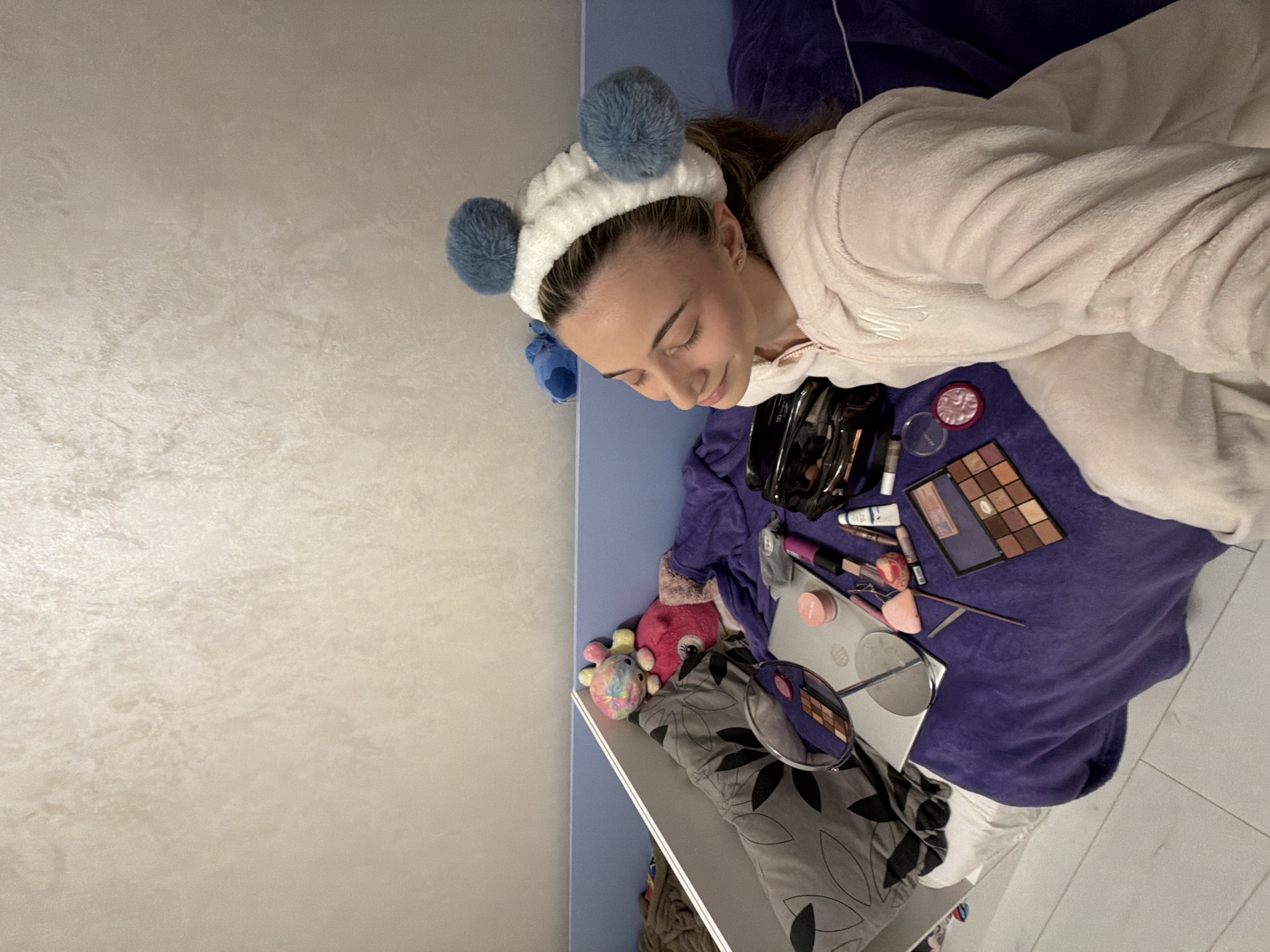 A young female is seated indoors on a bed covered by a purple fluffy blanket. She wears a light pink, fuzzy zippered loungewear suit featuring an embroidered initial on the left chest area, and a white fluffy headband with two prominent blue pom-poms. Her eyes are closed, and her head is tilted slightly to the right with a faint smile. Her left ear displays a small stud earring.
To her left, a grey pillow with black leaf-like patterns is present. Partially visible near the pillow are a colorful plush octopus and a pink plush animal. A blue plush toy is visible on a blue bed frame behind her head. A silver laptop, closed, rests on the purple blanket, surrounded by various makeup items. These include a silver-framed round magnifying mirror on a stand, an eyeshadow palette containing numerous brown and neutral shades, several makeup brushes, a pink compact, various tubes of lip products, and a black cosmetic bag. The wall behind the bed features a light beige, textured finish. Light-colored laminate flooring is visible in the lower left portion of the scene. The precise location within the city of Plovdiv, Bulgaria, is not discernible from the image.