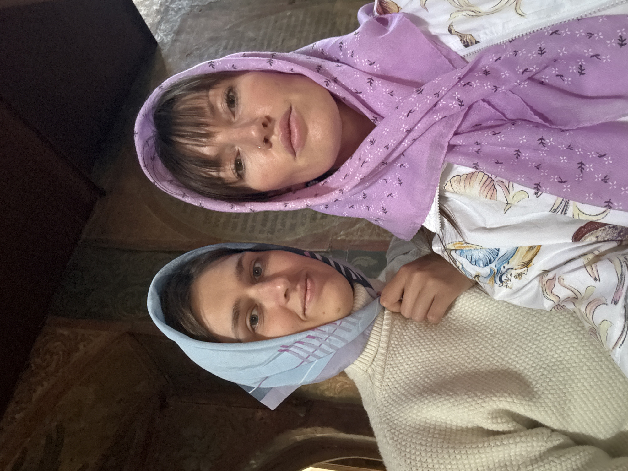 Two women, wearing headscarves, are pictured in what appears to be an interior space. The woman on the left has a light blue headscarf and is wearing a cream-colored sweater. The woman on the right has a purple headscarf and is wearing a white jacket with a floral pattern. The background features textured walls and architectural elements that suggest an older building, potentially a place of worship or historical significance, possibly in Moscow, Russia. The lighting is dim, creating a warm atmosphere.