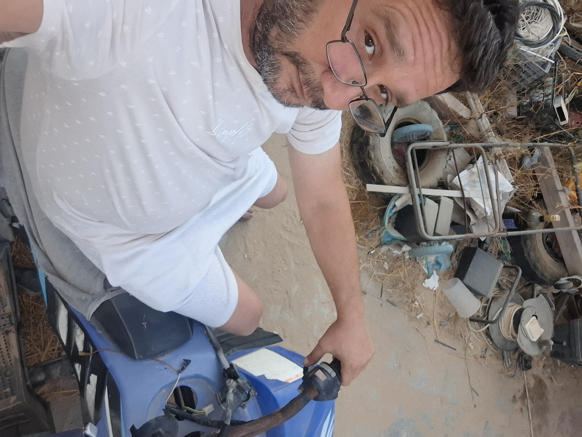 The image depicts a close-up, first-person view of a man sitting on a blue ATV. The man is wearing a light-colored t-shirt with a subtle pattern of small white sailboats and white shorts. He has short, dark hair, a beard, and is wearing glasses. His right hand is on the handlebar of the ATV. The background is cluttered with various objects, including tires, metal frames, plastic containers, coiled rope, and dried vegetation, suggesting an outdoor storage or work area. The ground appears to be dirt or sand. The scene is likely in Yerocham, Israel, given the context provided.