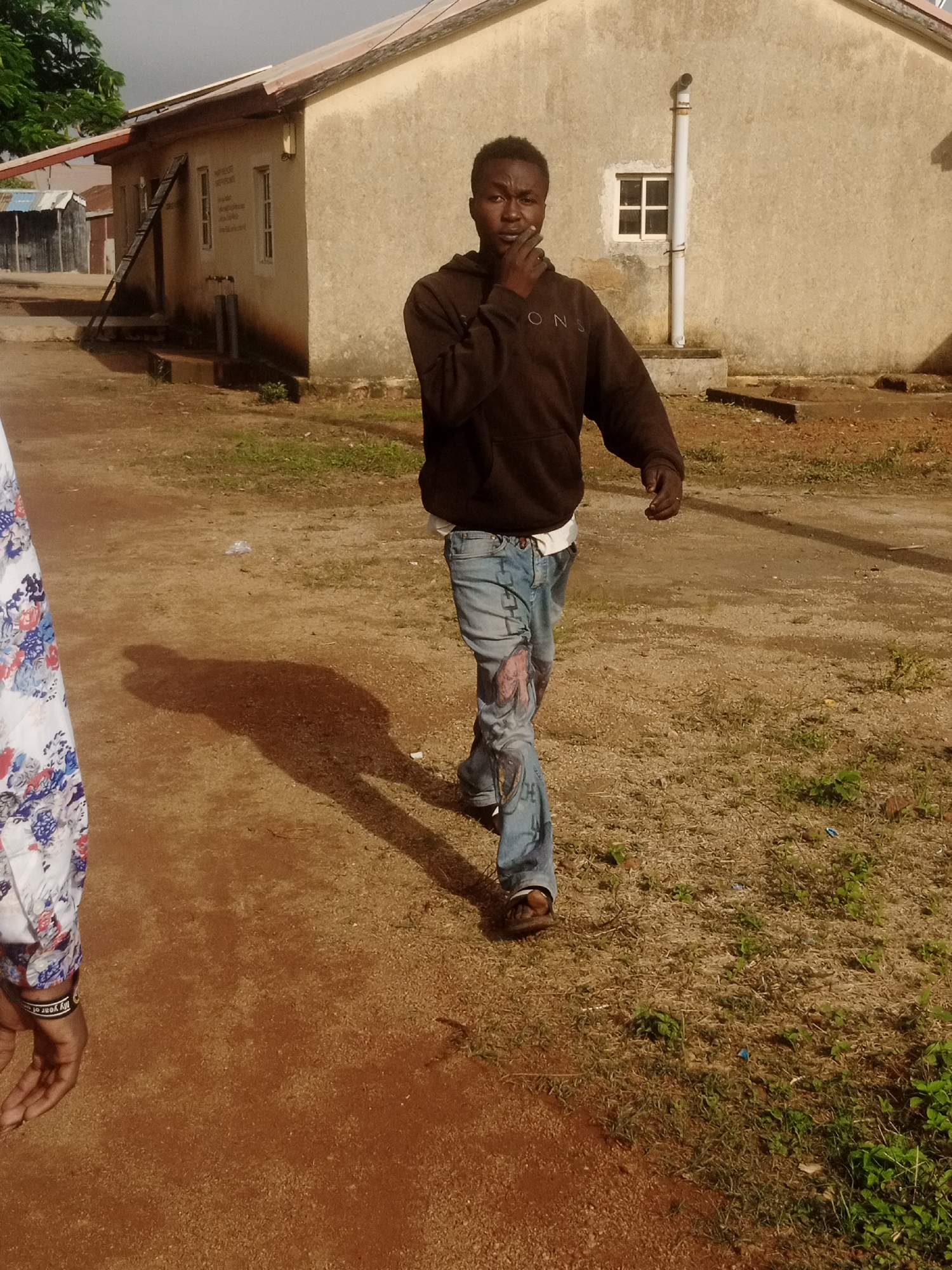 A young Black male, dressed in a dark brown hooded sweatshirt and distressed denim jeans with colorful paint or fabric patches, walks towards the camera on a dirt path. The man has his hand to his chin, appearing contemplative or mid-sentence. A woman, partially visible on the left, wears a white top with a floral pattern and a black bracelet. They are on the grounds of a weathered, pale yellow building with a corrugated metal roof and a single window visible. A ladder leans against the building, suggesting ongoing construction or maintenance. The environment is dry and somewhat arid, with patches of dry grass and sparse green vegetation. The lighting suggests it is daytime.