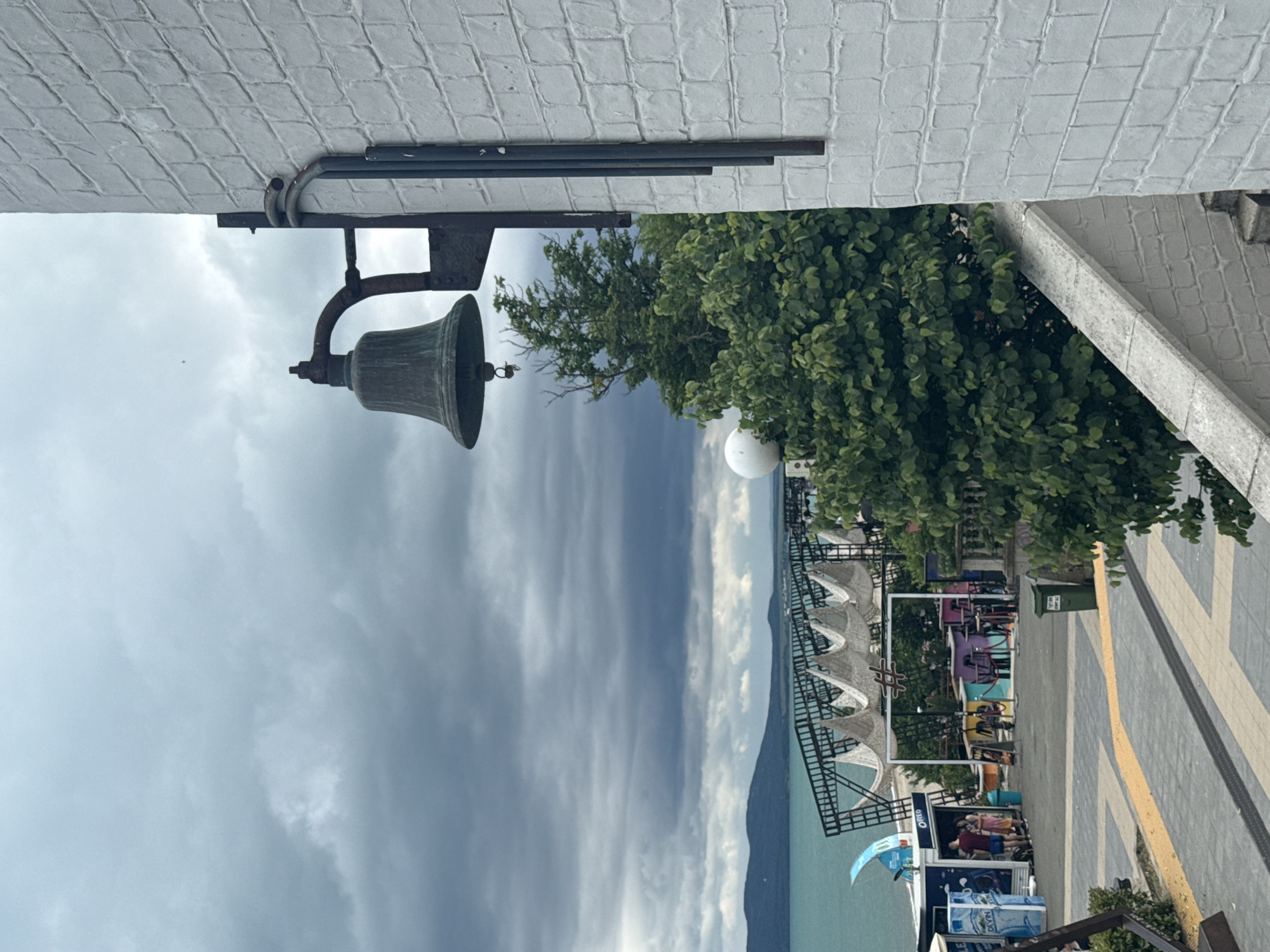 The image depicts a view from a raised walkway or staircase overlooking a coastal promenade in Burgas, Bulgaria. In the foreground, a white brick wall with a rough texture runs along the right side of the frame. Mounted on this wall is an old, dark-colored bell with a rusty bracket. Below the bell, a dense green tree occupies the middle ground, partially obscuring a white spherical object and a black structure extending from the wall.
Beyond the tree, the promenade is visible, paved with gray and yellow tiles. Several small kiosks or stalls line the promenade, one of which displays the "OREO" logo. A few people are standing near the stalls. Further back, a black metal structure supports a series of white, tent-like awnings. A large body of turquoise water, likely the Black Sea, stretches to the horizon, with a dark, hilly coastline in the distance. The sky is overcast with heavy, dark clouds. The overall atmosphere is that of an urban seaside area on a cloudy day.