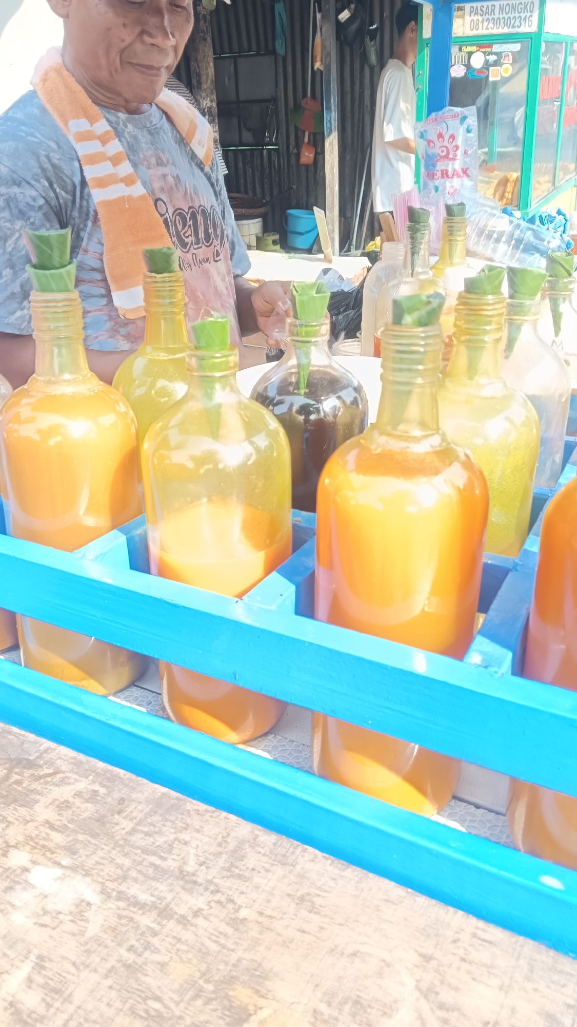 The image depicts a street vendor in Surakarta, Indonesia, likely at a local market or roadside stall. The vendor, an older individual wearing a grey t-shirt and an orange and white striped towel around their neck, is partially visible on the left side of the frame. In front of the vendor, a display of glass bottles filled with brightly colored liquids is prominently featured. The bottles are arranged in a blue wooden crate. The liquids appear to be a vibrant orange, possibly fruit juices or traditional beverages. Some bottles have green leaf-like stoppers. In the background, a partially obscured person in a white shirt is visible near a stall with signage. The environment appears to be outdoors, with corrugated metal sheeting and a wooden structure suggesting a basic, functional setup. The overall impression is a scene of local commerce in a tropical or subtropical setting, characterized by the bright sunlight and the colorful display of beverages.