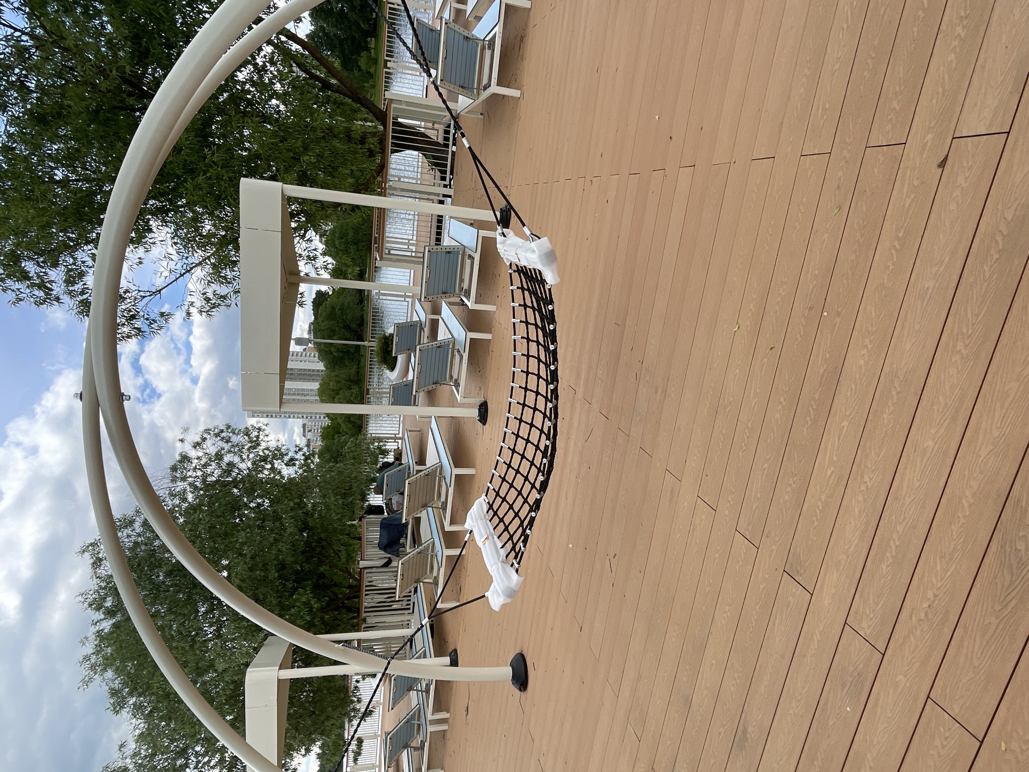 The image shows a wooden deck with a series of lounge chairs and a net swing. The swing is wrapped in white protective material. The deck is located near a park with trees and a building in the background. The location is Moscow, Russia. Several lounge chairs are located to the left.