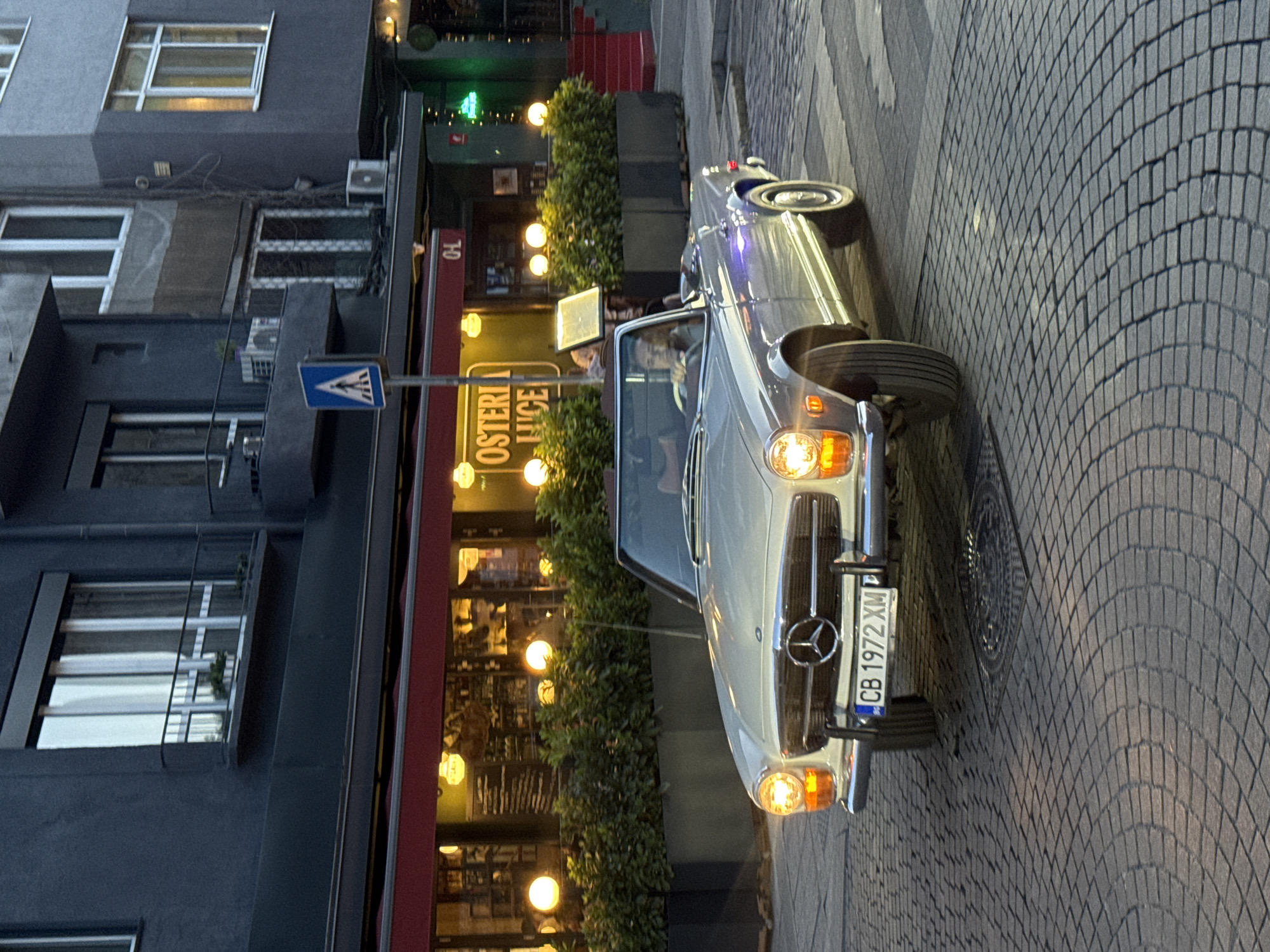 A vintage silver Mercedes-Benz convertible, license plate "CB 1972 XM," is driven on a cobblestone street in Sofia, Bulgaria. The car is illuminated by its own headlights and the warm glow of streetlights. Behind the car, a building features a restaurant with outdoor seating, indicated by potted plants and glowing spherical lights. The building's facade is dark, with visible windows and balconies. A blue pedestrian crossing sign is affixed to a pole in front of the restaurant. The overall atmosphere suggests a late evening or dusk setting in an urban environment.