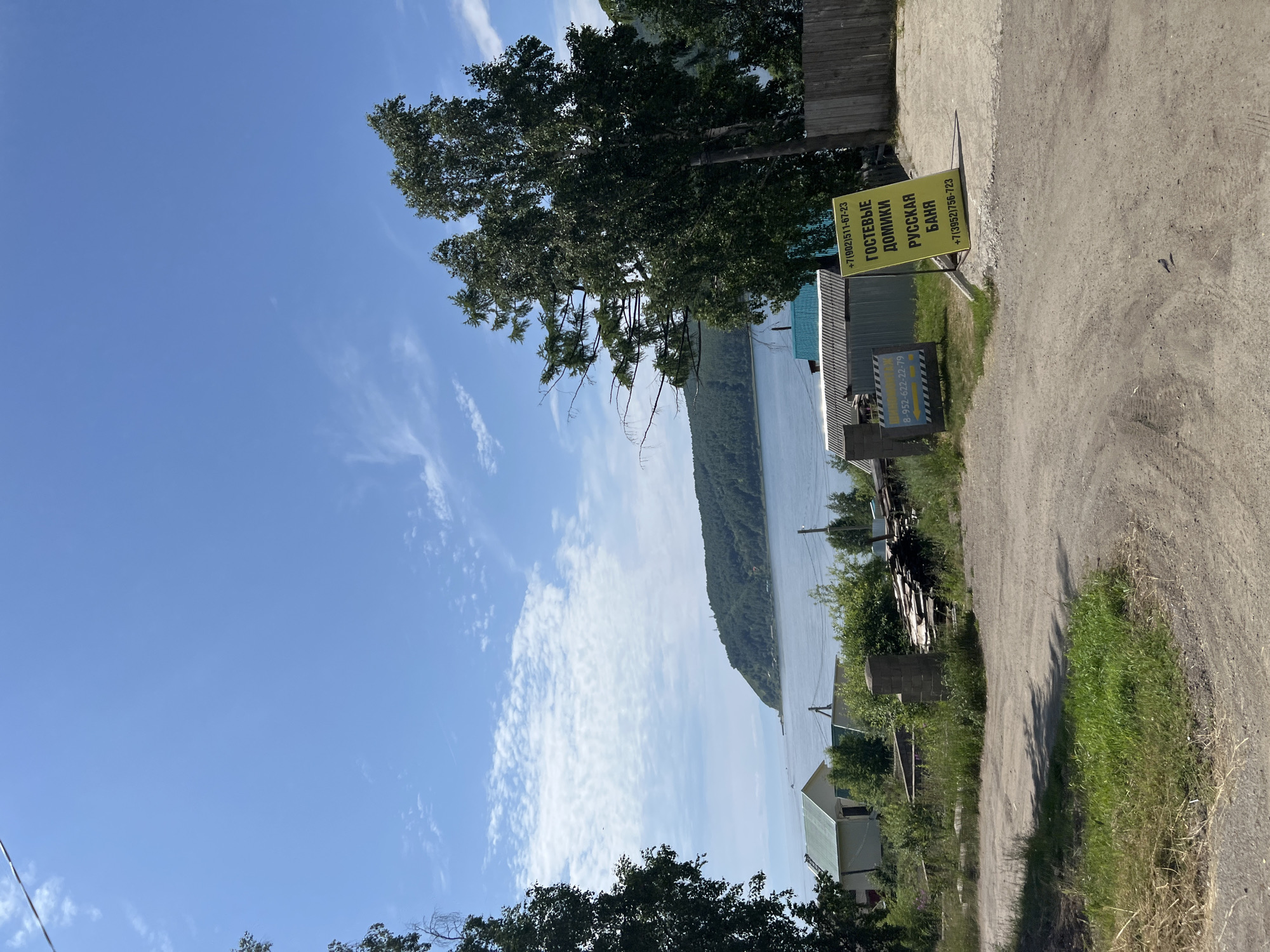 The image depicts a sunny day scene in Listvyanka, Irkutsk Oblast, Russia, likely near the shores of Lake Baikal. The foreground features a dirt road or pathway with sparse green grass and dry straw on the sides. To the right of the road, a yellow sign with black text is prominently displayed, advertising "Guest Houses" and "Russian Bathhouse" with phone numbers. A wooden fence borders the right edge of the scene. Behind the road, several low-rise buildings with light-colored walls and green or turquoise roofs are visible, nestled amongst lush green trees. In the mid-ground, the vast expanse of a large body of water, presumably Lake Baikal, stretches towards a densely forested headland in the distance. The sky is a vibrant blue with scattered white clouds, indicative of fair weather. The overall impression is of a rural or semi-rural location, possibly a holiday or tourist area, with a focus on natural scenery and outdoor activities. There are no people visible in the image.