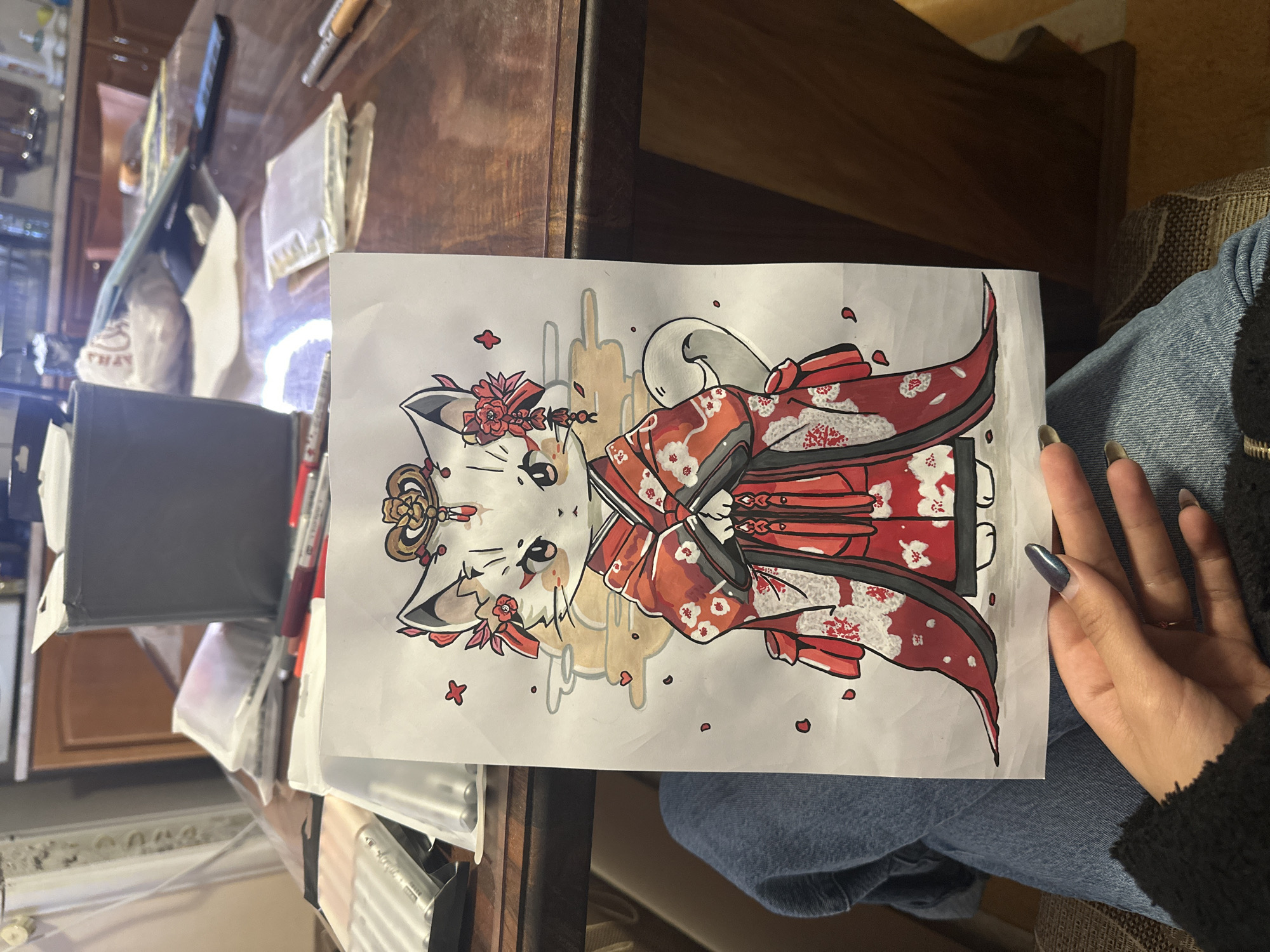 A person's hand holds a drawing of a stylized cat in a red and white kimono. The drawing is on white paper, and the cat figure is surrounded by grey and brown decorations. The person is wearing blue jeans and a black sweater. The scene is inside a room with a wooden desk and cabinets. Writing and drawing supplies are on the desk. The room's exact location in Burgas, Bulgaria, cannot be determined from the image.