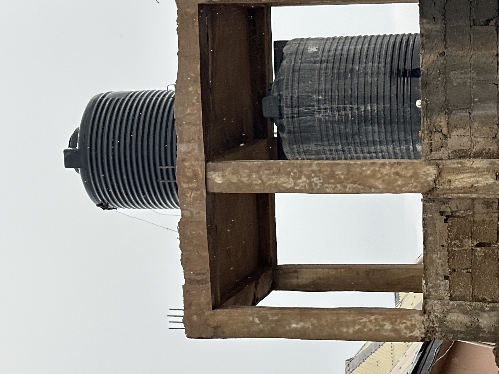 This media file features a close-up view of two large, black, ribbed plastic water tanks. They appear to be positioned within a partially constructed or deteriorated concrete and brick structure. The structure has rough, unfinished concrete beams and a wall made of concrete blocks with visible mortar. The tanks are cylindrical and have a corrugated surface. One tank is positioned on the left, and another is visible to the right, partially obscured by the structure. The background is a bright, overcast sky, suggesting daytime with diffused light.
The scene seems to depict a water storage or supply system, possibly in a construction or maintenance phase, given the unfinished nature of the surrounding structure. There are small white specks scattered across the image, which could be dust or debris. On the left side of the frame, some thin metal rods or wires are protruding from the concrete structure. The image's orientation is rotated, making the sky appear on the left and the ground or lower structures at the bottom. No people are visible, and the overall mood is neutral, focusing on the objects and their environment. The location context provided is Jalingo, Nigeria. There is no discernible text in the image.