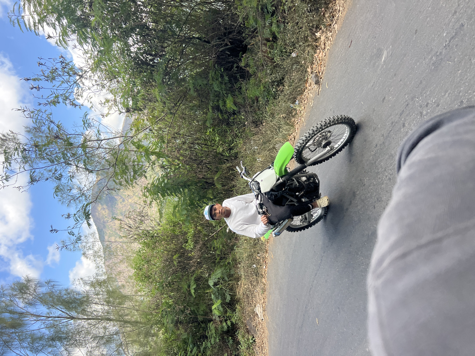 A young man is riding a dirt bike on a paved road. He is wearing a white long-sleeved shirt, black pants, and a blue and white baseball cap. He has on white clogs. The dirt bike is green and white. Behind the man, there is a lush green hillside with trees and vegetation. In the distance, a mountain can be seen under a blue sky with white clouds. The scene appears to be in Kintamani, Indonesia, known for its volcanic landscapes and natural beauty.