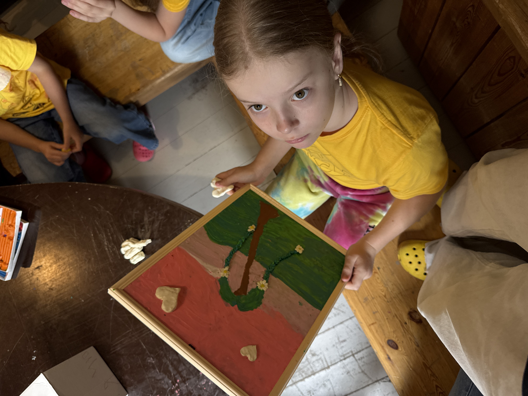 A high-angle medium shot captures a young girl wearing a yellow t-shirt and tie-dye pants, sitting at a wooden table and holding a small framed artwork. The artwork depicts a tree with green foliage, a brown trunk, and two light-colored hearts on a pink and tan background. The girl’s left hand is holding the artwork, and her right hand is holding a small piece of light-colored material. Her gaze is directed upwards towards the camera. In the background, the edge of another wooden table is visible, with some art supplies, including a box of orange modeling clay, on it. Another child, wearing a yellow t-shirt and blue jeans, is seated on a wooden bench in the background. The floor is composed of light-colored planks. The image is likely taken indoors, in a setting that suggests a crafting activity. The scene does not provide any specific geographical indicators of Moscow, Russia.