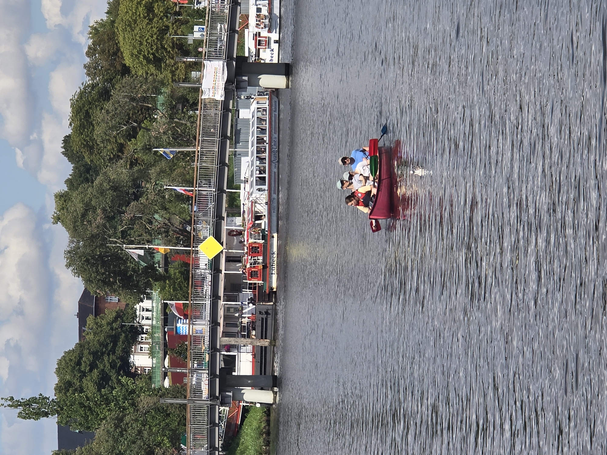 This is a photograph of a red canoe with four people in it on a river in Lübeck, Germany. The canoe is in the foreground of the image, with the people inside paddling. In the background, there is a bridge with cars and people on it. There are also buildings and trees on the shore. The sky is blue with some clouds. The river is wide and calm, with ripples on the surface. The water is dark blue. There is a ferryboat in the background with passengers. The bridge is made of metal and concrete. The buildings in the background are red brick and white. The trees are green and leafy. The sky is blue with white clouds. The overall mood of the image is peaceful and serene.
