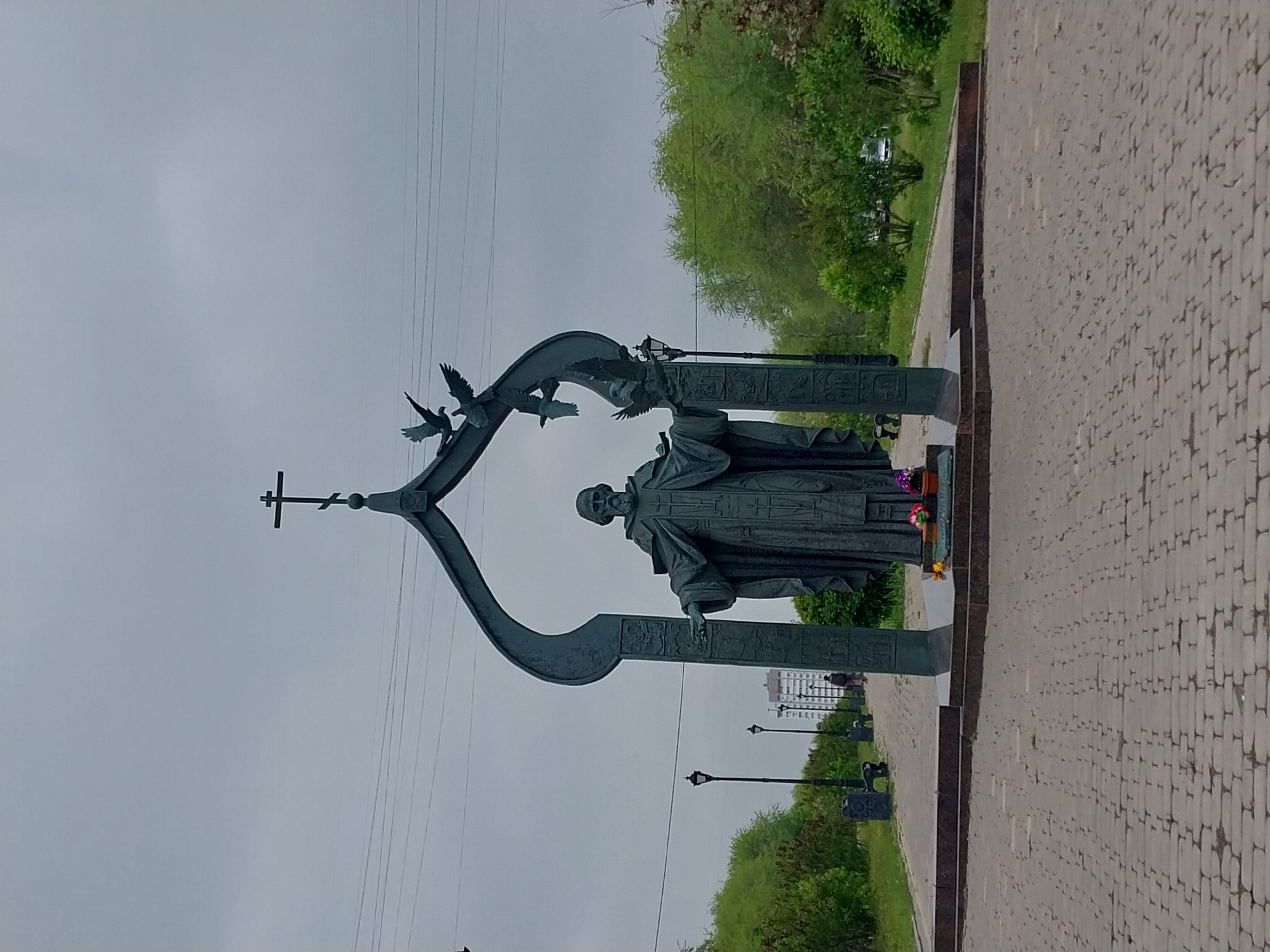 The image shows the monument to St. Innocent of Kherson in Orenburg, Russia. The statue depicts a man in ecclesiastical robes. It is framed by a dark metal arch with a cross at the top and three stylized doves in flight. The base is constructed of a reddish stone. Flowers are positioned at the base of the statue. The surrounding environment includes a paved walkway, trees, street lamps, and a building in the distance. The sky is overcast.