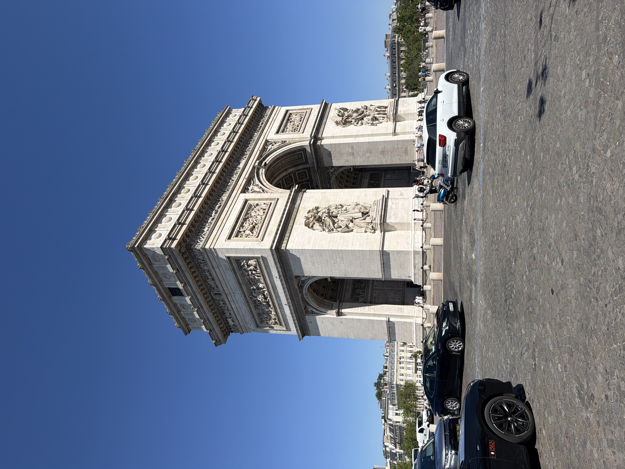 The image depicts the Arc de Triomphe in Paris, France, under a clear blue sky. The monument is constructed from light-colored stone with detailed carvings. A cobblestone plaza surrounds the Arc de Triomphe. Several vehicles are parked in the plaza, including a white SUV and multiple dark-colored cars. Small groups of people are visible along the edge of the plaza, near the monument. Buildings and trees are seen in the background, indicating an urban setting.