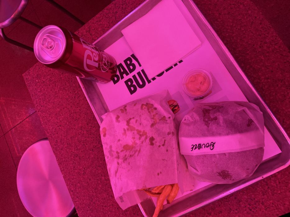 A tray holds a meal: a Dr. Pepper can, a small container of sauce, a burger, and fries wrapped in paper. "BABY BURGER" is printed on the tray's liner with a white napkin resting near the top. The burger, also wrapped in paper, has a label reading "Bourke." The meal sits on a table next to a modern chair with a circular seat. The image is bathed in red lighting.