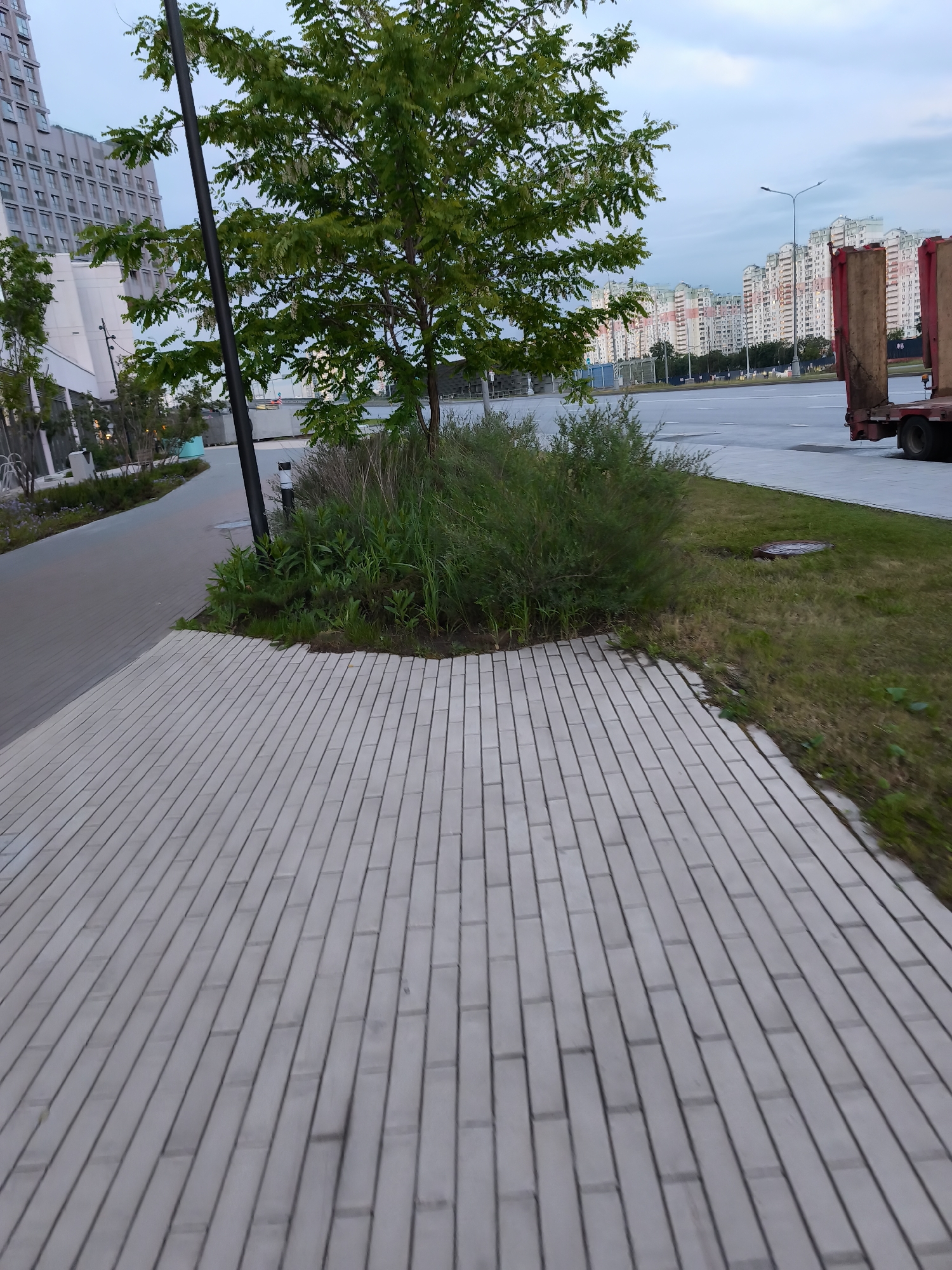 The image shows a view of an urban landscape in Moscow, Russia. In the foreground, there is a paved brick pathway, leading to an area with a small tree, shrubbery, and a light pole. To the left of the pathway is a narrower pedestrian path. In the background, across a broad road, are several tall residential apartment buildings. To the right of the image, there is a patch of grass and a portion of a truck. The sky is overcast.