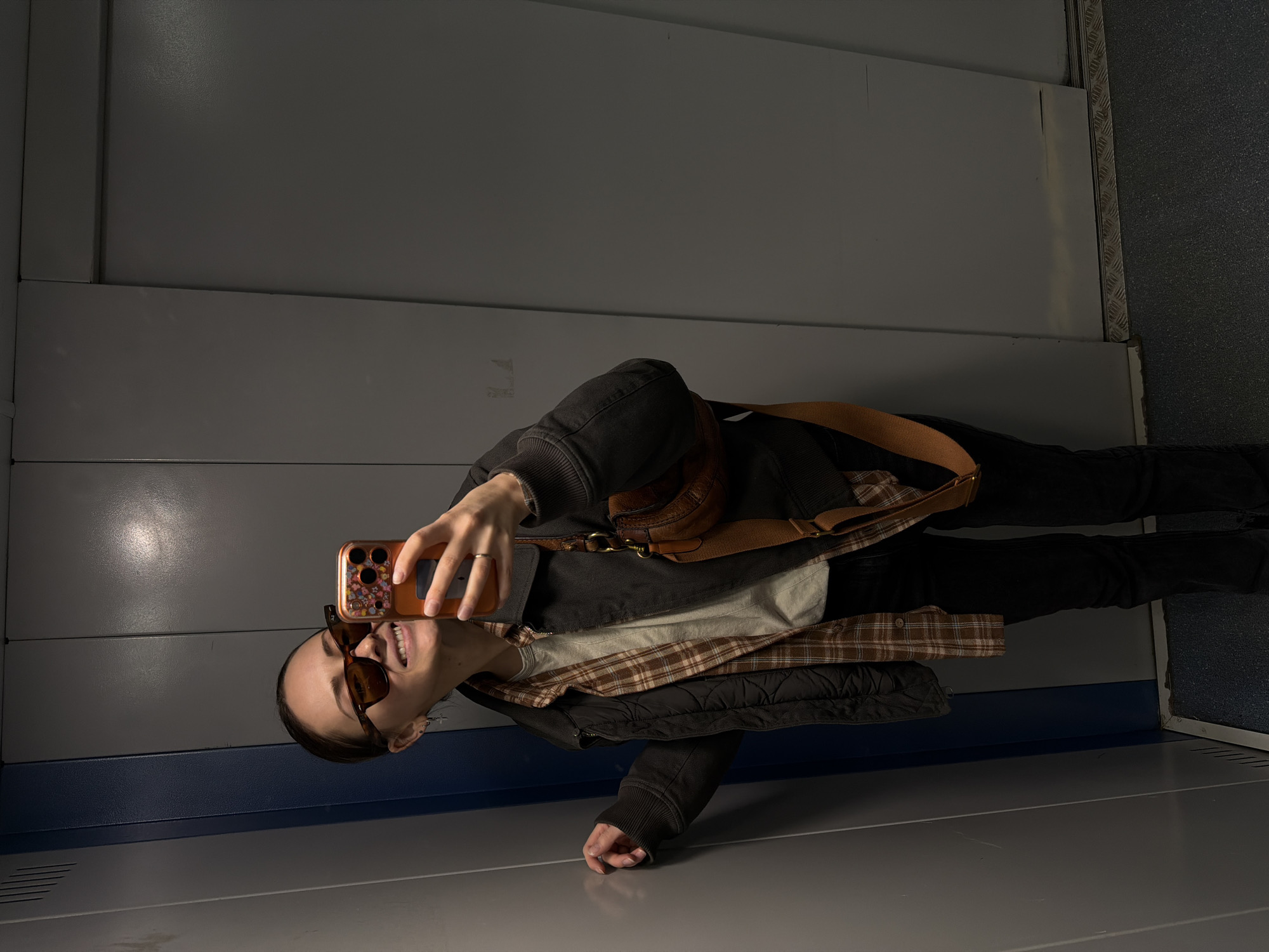 A young woman stands inside an elevator in Khimki, Russia, taking a selfie. She is wearing sunglasses, a light-colored shirt, a brown plaid flannel, a black jacket, and black pants. A brown purse with a tan strap is slung across her body. Her right hand holds a smartphone with a decorative case. The elevator walls are gray with blue trim. The floor is gray.
