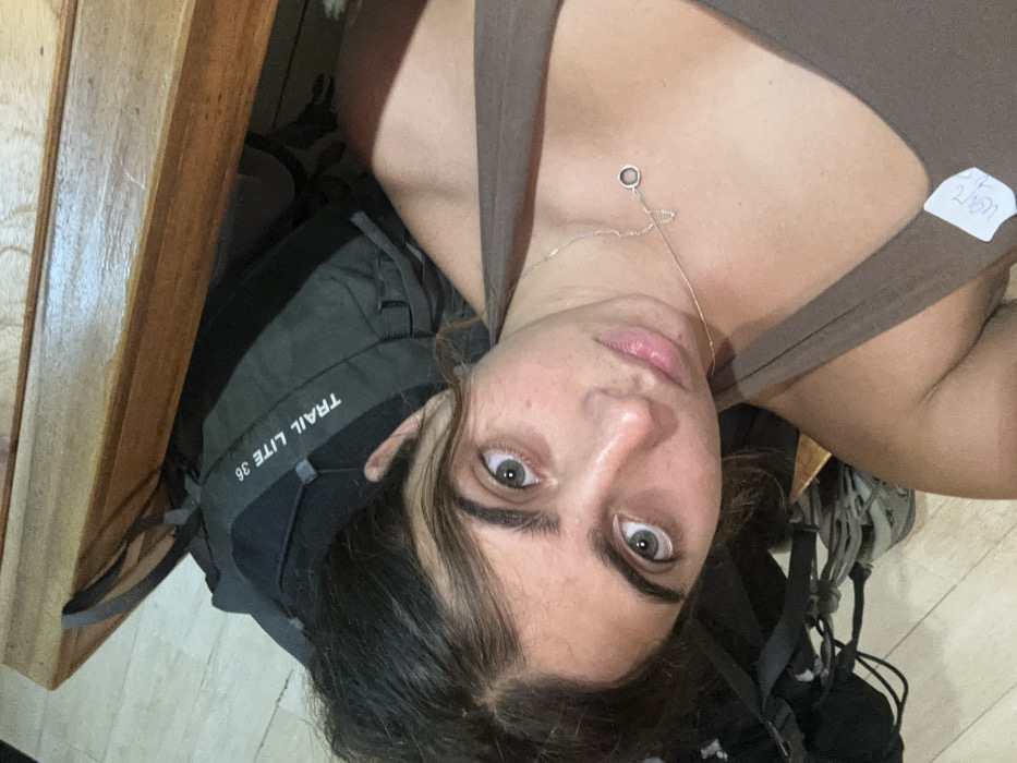 The image displays an indoor scene within Surat Thani City Municipality, Thailand. A light-skinned young adult female, with dark hair, prominent eyebrows, and light-colored eyes, is centrally featured, looking directly at the camera. She is wearing a brown tank top and a thin silver necklace with a small circular pendant. A white sticker with partially legible writing, possibly "200 Baht," is affixed to the front of her tank top. Positioned behind her is a dark grey backpack, identified by the text "TRAIL LITE 36" printed vertically on its side. A light-colored tiled floor is visible in the lower left, and a wooden surface, likely part of a bed frame or furniture, appears in the upper portion of the frame. The lighting indicates an interior space, consistent with an accommodation setting.