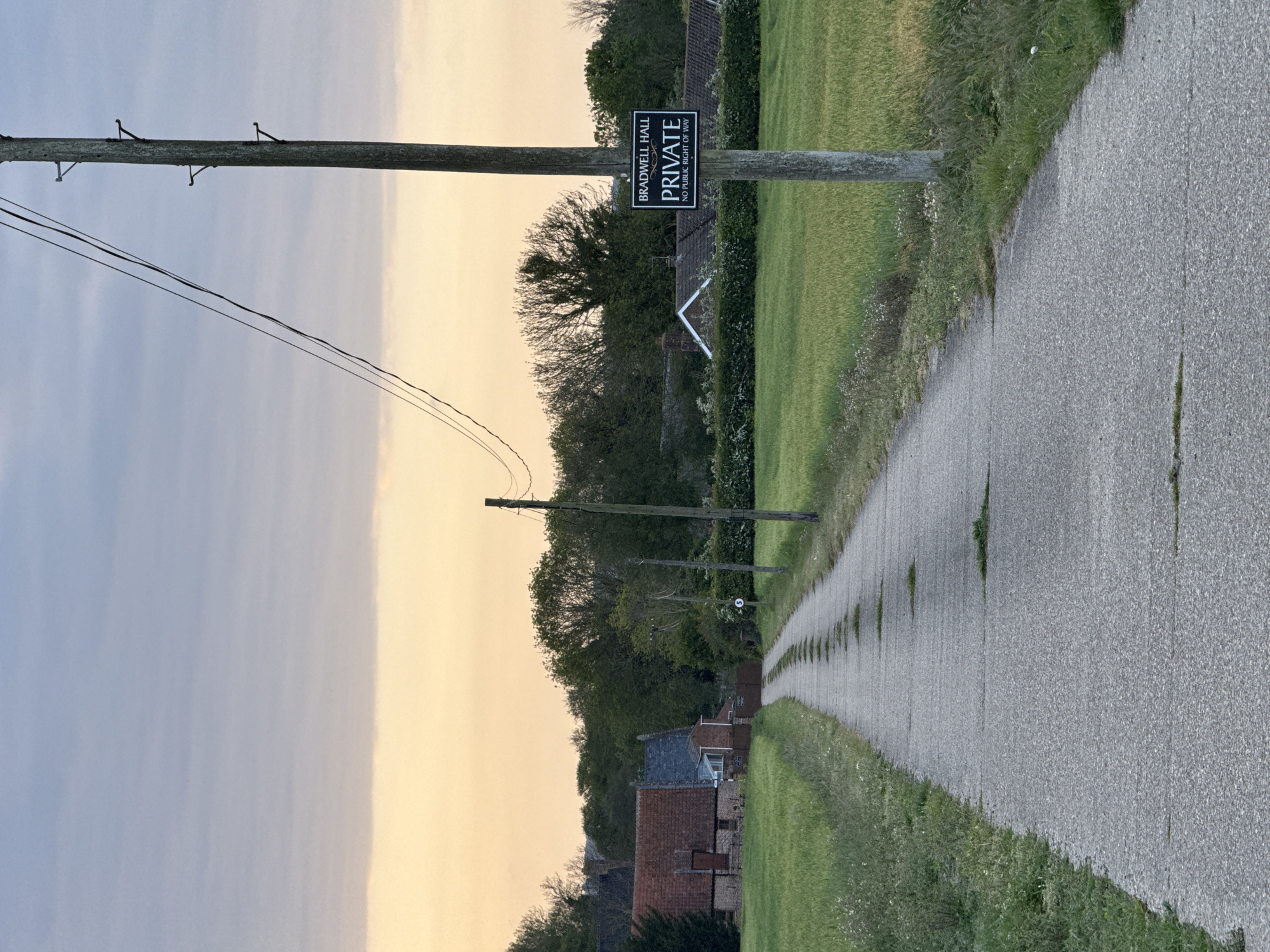 A narrow, straight paved road extends into the distance, framed by grassy verges on either side. A sign on a wooden post reads "BRADWELL HALL PRIVATE NO PUBLIC RIGHT OF WAY". Utility poles line the road, carrying power lines. In the background, trees and rooftops of houses are visible. The sky is divided into light blue and orange sections. The image likely depicts a rural or suburban area near Great Yarmouth, United Kingdom.