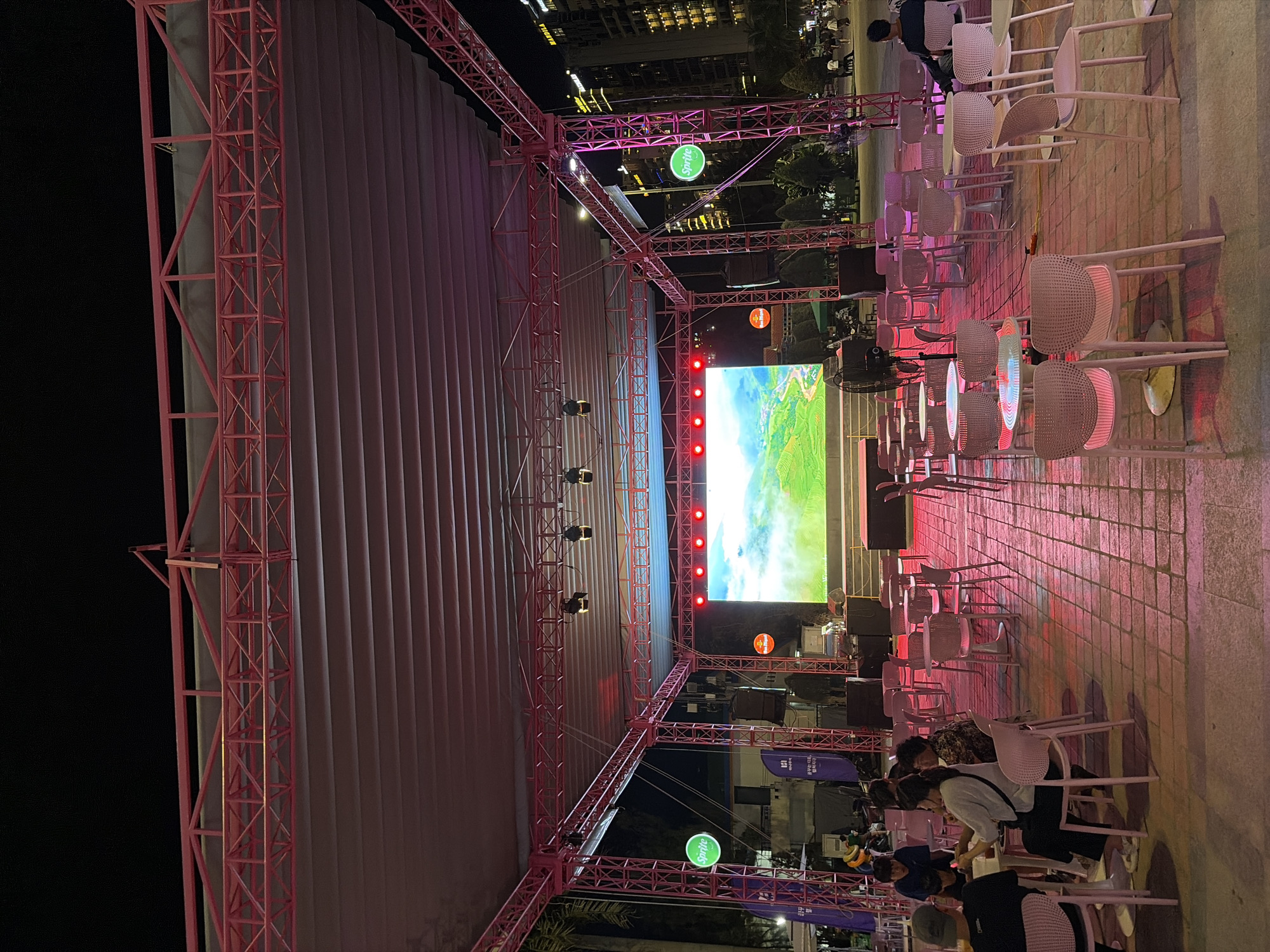 This image captures an outdoor evening scene in Đà Nẵng, Vietnam, likely at a public square or entertainment venue. A large, illuminated stage with a screen displaying a landscape scene is the central focus. The stage is framed by a pink metal truss structure that supports a white canopy overhead. Pink and red lights cast a colorful glow on the ground, which is paved with square tiles.
In the foreground, several groups of people are seated at tables and chairs. Some are engaged in conversation, while others appear to be observing the stage. The tables are small and round, and the chairs are modern, light-colored plastic. The overall atmosphere suggests a casual gathering or event.
Several branded signs are visible, including a green circular sign with the "Sprite" logo on the left side of the stage. In the background, a tall building with illuminated windows can be seen, indicating an urban setting. Trees and other vegetation are present, suggesting a park-like or landscaped area surrounding the venue. The night sky is dark, with the lighting of the event and the city providing illumination.