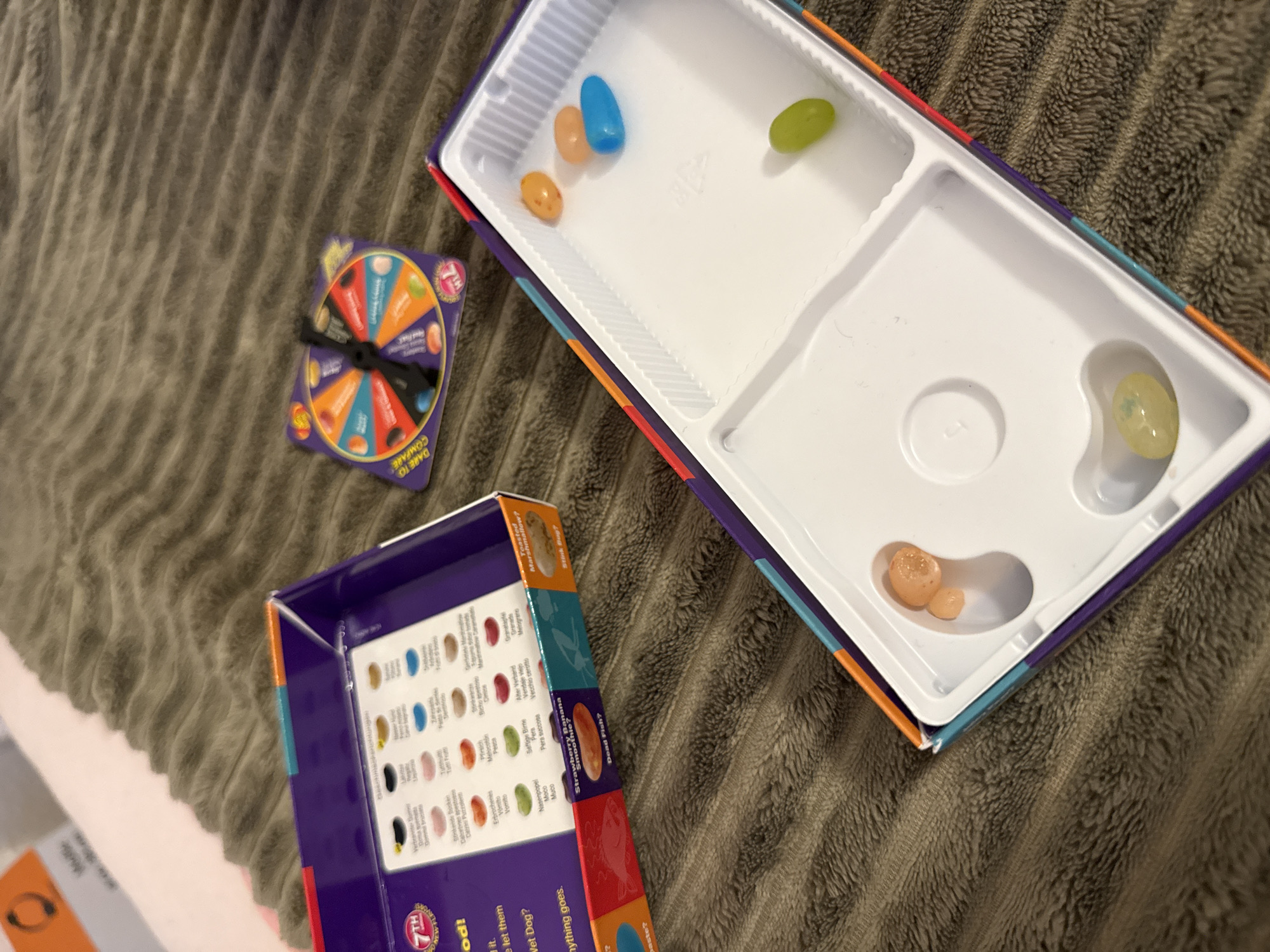 The image shows a Jelly Belly Beanboozled game set. The game box is purple with colorful edges, and the interior tray contains several jelly beans. There are orange, blue, and green jelly beans scattered in the tray. A spinning wheel with different colored sections and images of jelly beans is also visible, along with the question "DARING TO COMPARE". The game appears to be set on a brown, textured surface, possibly a blanket or rug. The scene does not provide any information about the city of Bruchköbel, Germany.