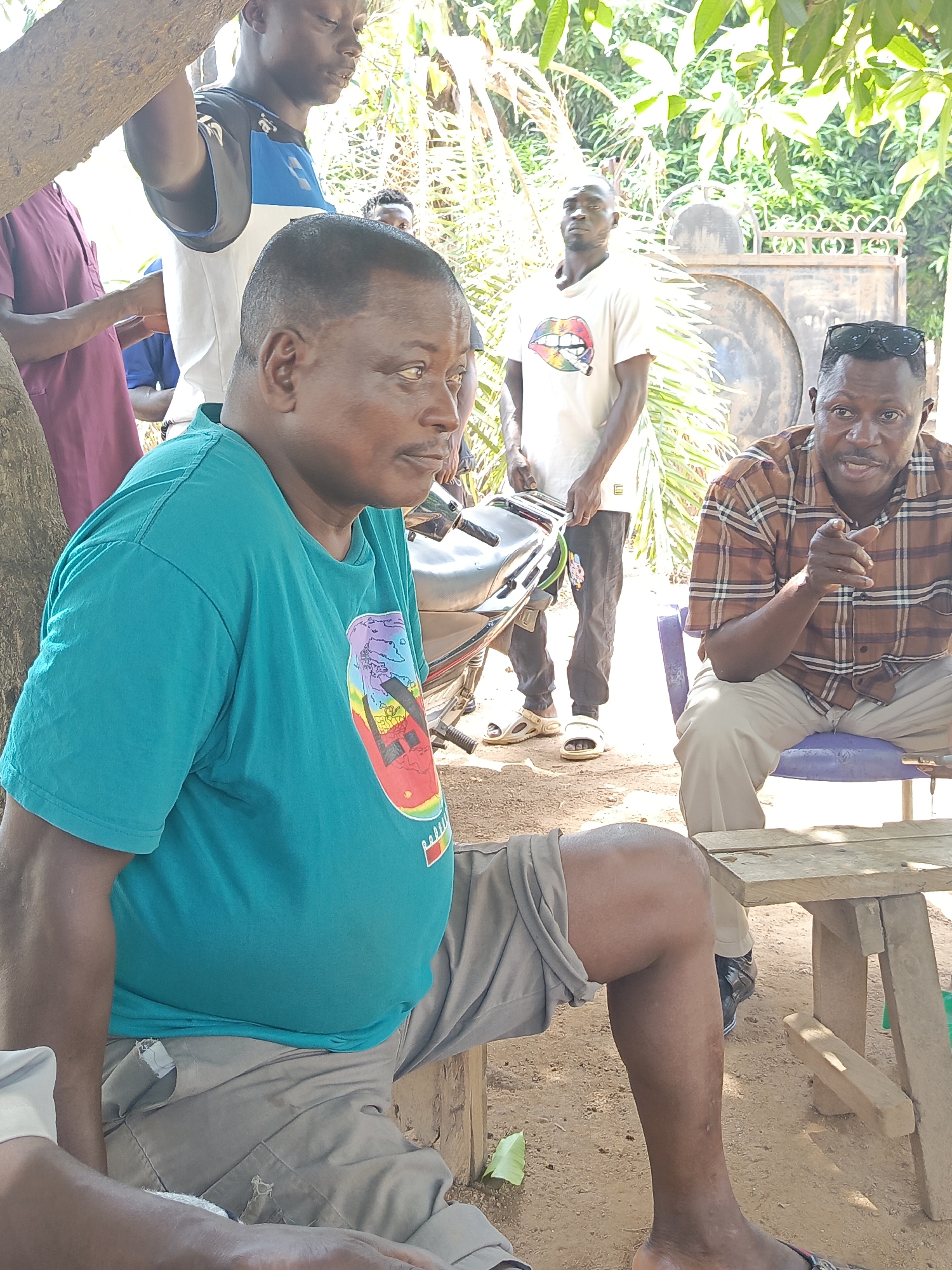 The image captures an outdoor scene in Akwanga, Nigeria, featuring a group of men. In the foreground, a man wearing a teal t-shirt and grey shorts sits on a makeshift wooden seat. Beside him, another man in a brown plaid shirt and light-colored pants sits on a blue plastic chair, gesturing with his right hand. A small wooden table stands between them. Behind these two, other men are visible, including one wearing a white t-shirt with a graphic and another in a white and blue top. A motorcycle is parked behind the man in the teal shirt. The setting includes trees, foliage, and a metal gate in the background. The ground appears to be dirt. The time of day appears to be midday, considering the bright sunlight.