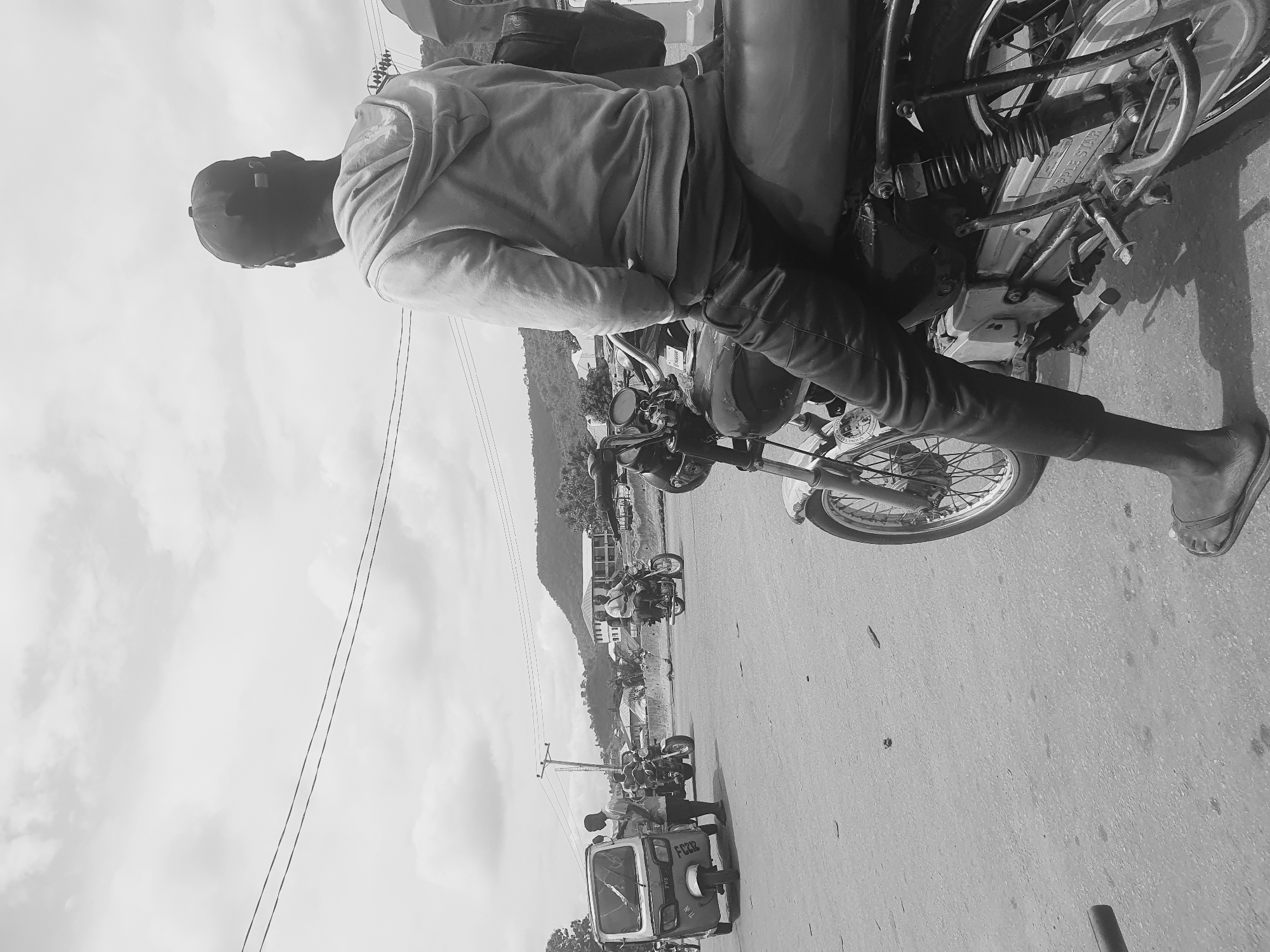 A person is sitting on a motorcycle on a paved road. The person is wearing a hoodie and jeans, and a cap. The motorcycle appears to be parked on the side of the road, with several other motorcycles and a "keke napep," or auto rickshaw, visible in the distance. The scene appears to be in an urban area of Lokoja, Nigeria. A mountain and buildings are visible in the background. Overhead power lines run parallel to the road.