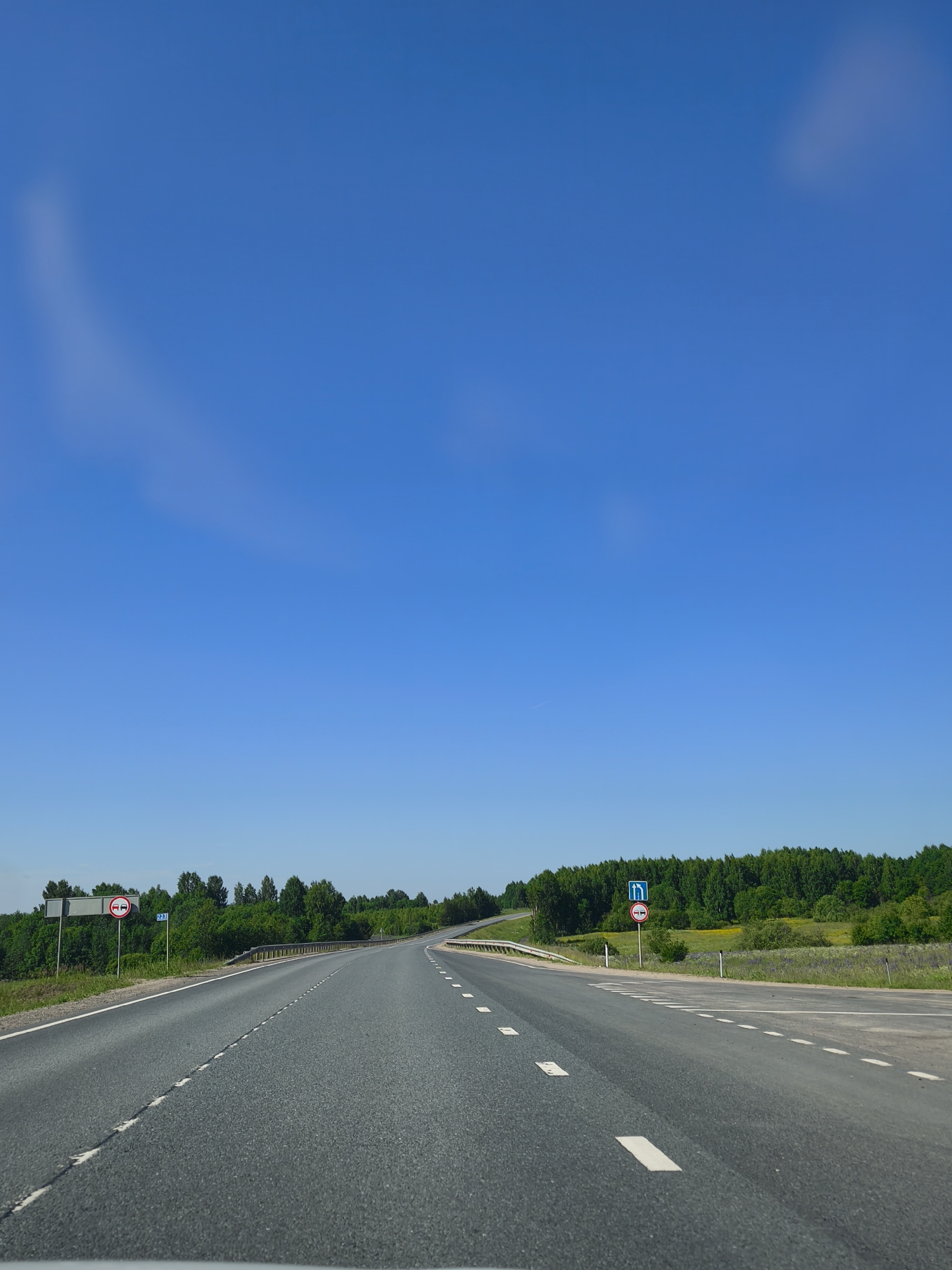 The image shows an asphalt road curving to the right, bordered by a metal guardrail on the left and a grassy shoulder with trees in the background. The sky is clear blue with faint, wispy clouds. There are two traffic signs visible on the left side of the road. The first sign, a circular red and white sign, depicts two cars side-by-side with a red bar through them, indicating no overtaking. The second sign, a blue square sign, shows a left turn arrow with a straight arrow to its right. The scene appears to be in a rural or suburban area, possibly on the outskirts of Makaryev, Russia, given the presence of trees and the general landscape. There are no people or vehicles visible in the immediate vicinity of the road.