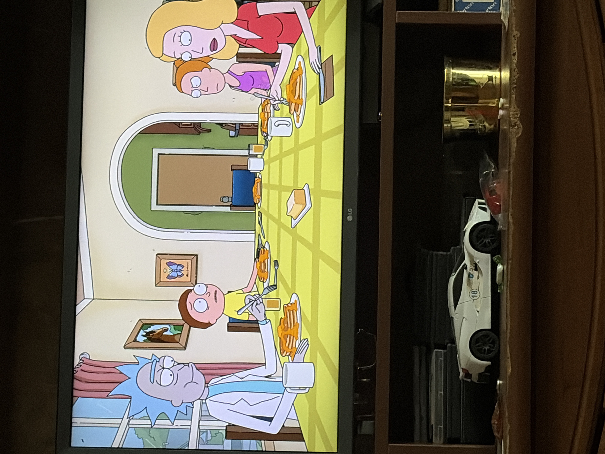The image displays a television screen showing an animated scene from the TV show Rick and Morty. The scene depicts Rick Sanchez, Morty Smith, Summer Smith, and Beth Smith sitting around a table, eating pancakes. The characters are animated in the show's characteristic style. The television set is situated on a wooden shelf unit, below which is a white toy sports car with the number 18 on its side and a golden cup. The background is dark, suggesting the room is dimly lit. The scene is set indoors in what appears to be a dining room.
