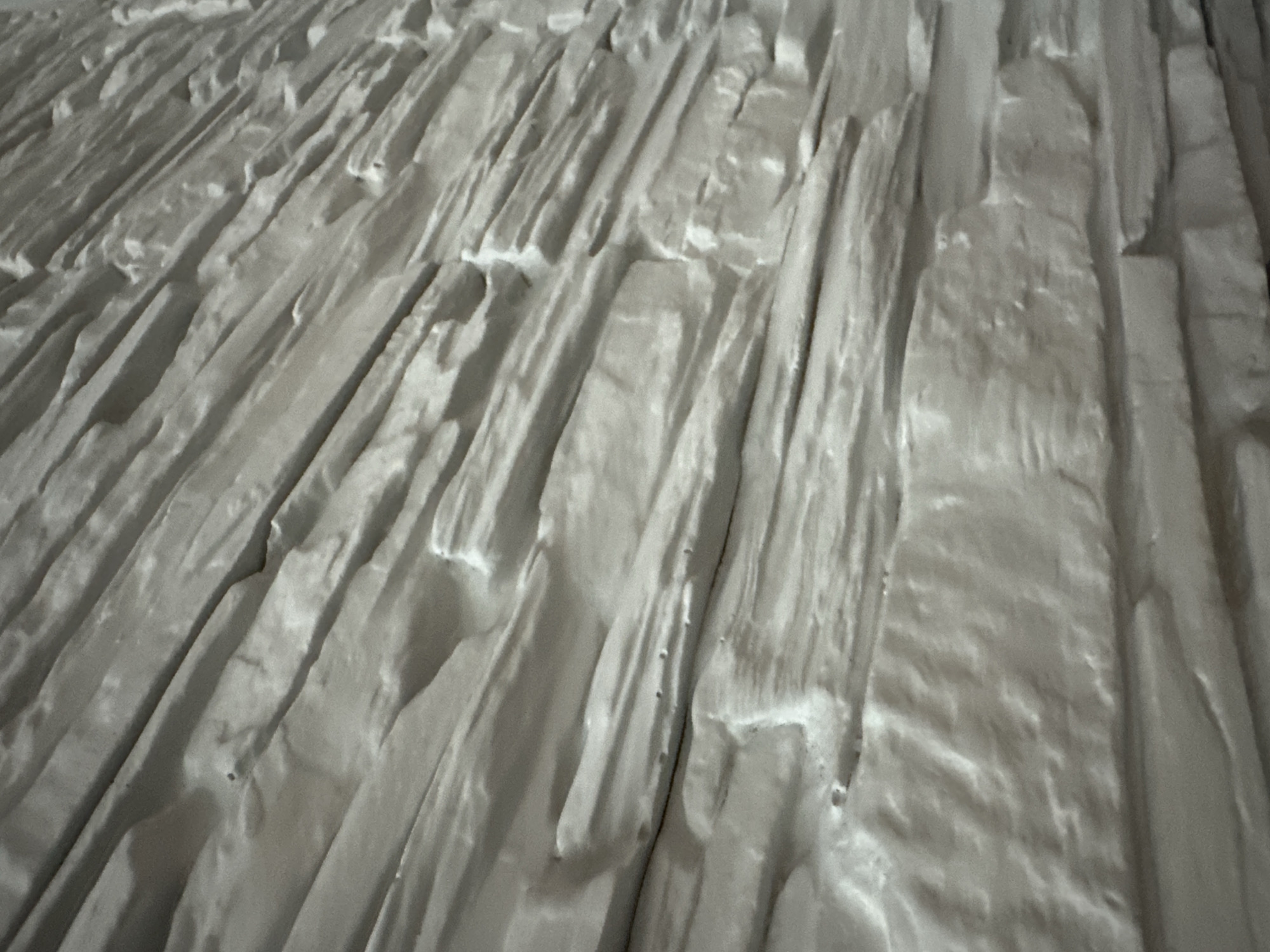 The image displays a close-up, high-angle view of a textured surface, devoid of people or discernible objects. The scene is abstract, showcasing a complex arrangement of elongated, irregular shapes that resemble planks, strips, or deeply grooved formations. These elements are predominantly oriented diagonally across the frame, running from the lower left towards the upper right.
The surface is rendered in shades of off-white, light gray, and creamy tones, with pronounced highlights and shadows that emphasize its three-dimensional relief. The texture is rough and uneven, characterized by numerous ridges, peaks, and deep valleys. The edges of these individual elements are often jagged or wavy, suggesting an organic, sculpted, or possibly weathered natural material rather than something uniformly manufactured. The lighting appears soft but directed, effectively illuminating the raised areas and casting shadows into the depressions, enhancing the sense of depth and form.
No specific activity or event is taking place; the image is a static portrayal of the surface's intricate structure. There are no indications of time of day, weather, or specific location cues. No text is visible. The overall impression is one of tactile richness and a robust, possibly mineral or handcrafted material.